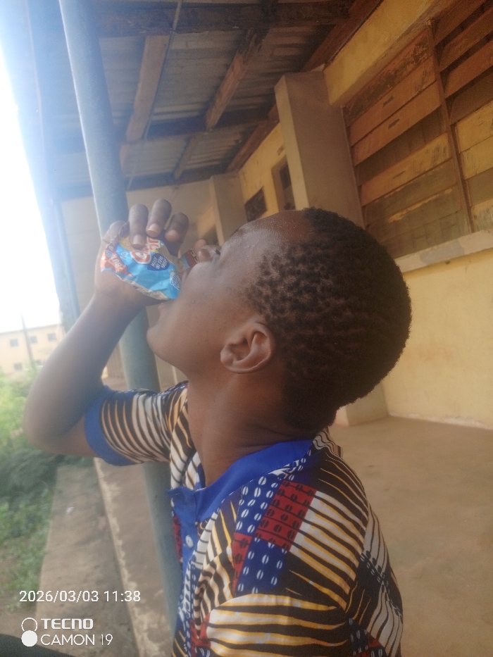 The image depicts an individual, observed from the right profile, actively drinking from a small, flexible beverage pouch. The individual has short, dark, closely cropped hair and wears a short-sleeved polo shirt with a solid blue collar and a distinct pattern featuring black, white, yellow, red, and blue geometric shapes and stripes. The right hand grips the multi-colored pouch, which displays red, blue, white, and yellow elements.
The scene is set in what appears to be a covered outdoor corridor or balcony, potentially part of a building in Onitsha, Nigeria. A prominent, thick, dark grey vertical metal pipe or structural column is visible in the left foreground. Overhead, exposed wooden rafters support a corrugated metallic roof. To the right of the subject, a light-colored building wall features two sets of horizontally slatted wooden windows, consistent with a louvered design. Beyond the covered area, on the far left, glimpses of green foliage and a segment of a multi-story building are visible under a bright, sunlit sky. The image bears a digital timestamp "2026/03/03 11:38" and "TECNO CAMON 19" in the lower-left corner.