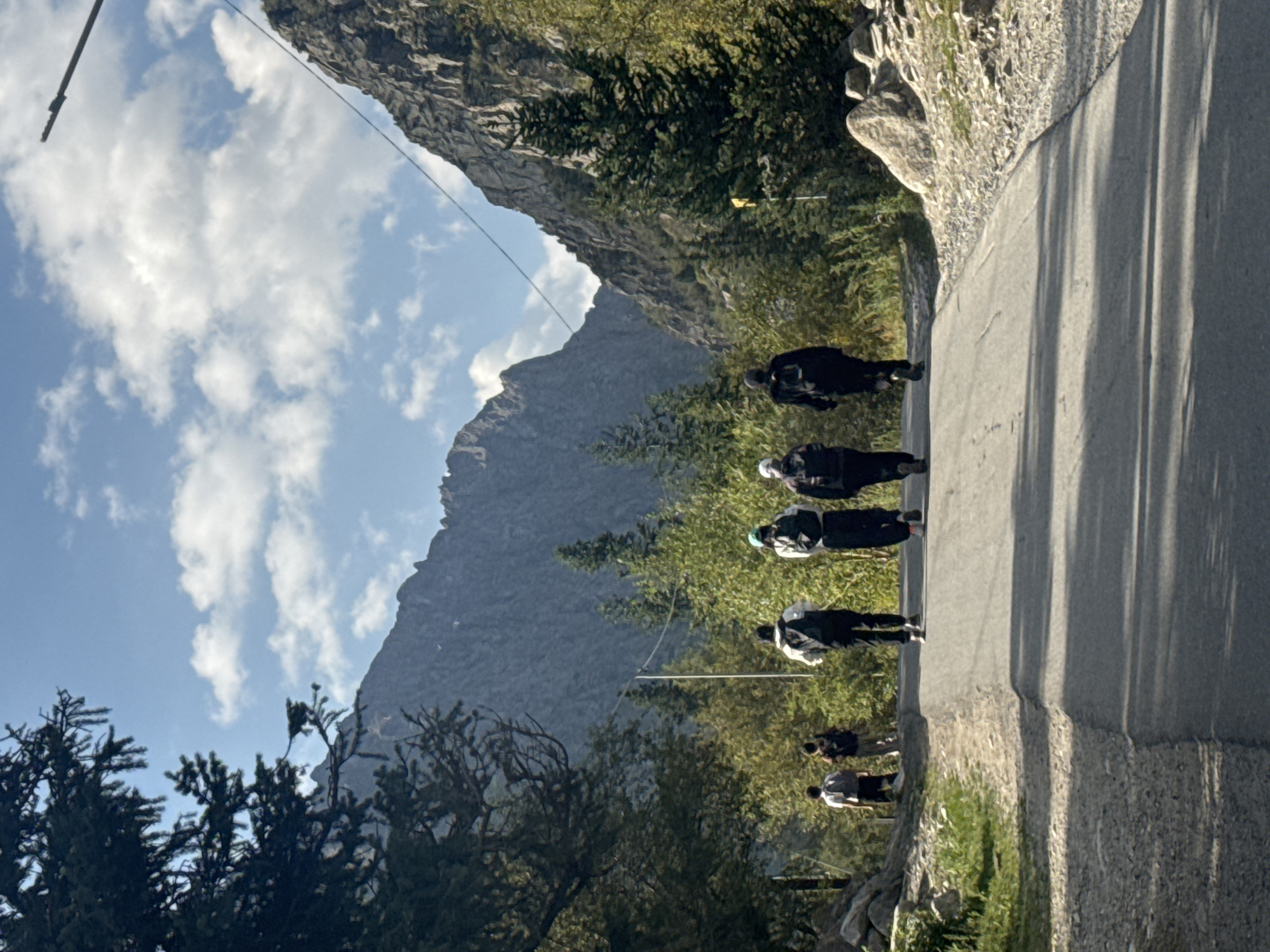 The image shows a group of five individuals walking away from the camera down a paved road, flanked by trees and rocky, mountainous terrain under a blue sky with scattered clouds. The individuals are dressed in casual outdoor clothing, and four of them are wearing backpacks. The group appears to be hiking or traveling in a remote, natural setting. The road leads into a valley between two large, steep mountains, suggesting a mountainous region. The vegetation consists of a mix of coniferous trees and deciduous trees, with some scrubby bushes along the roadside. The lighting indicates it is daytime, likely late morning or early afternoon. The presence of power lines strung across the valley suggests some level of infrastructure in the area.