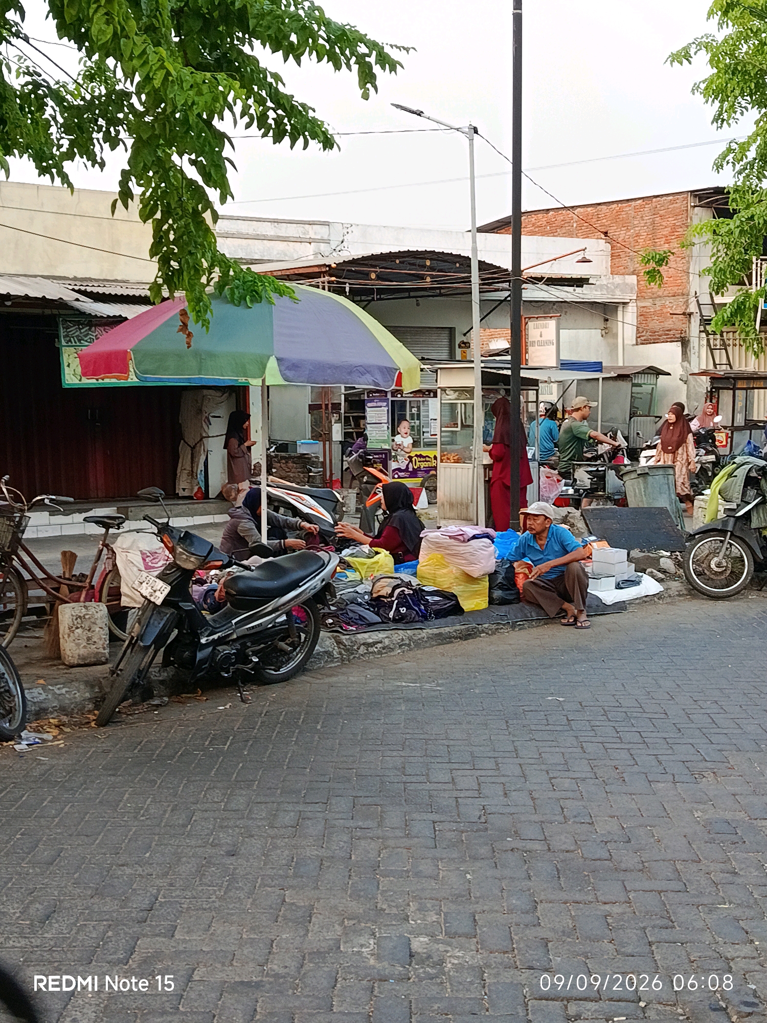 In Keboansikep, Indonesia, a bustling street scene unfolds with vendors and shoppers. A multi-colored umbrella shades a stall where several people are engaged in commerce. A motorcycle is parked near a bicycle, and various goods, including bags of clothes and other items, are displayed on the ground. Buildings with brick and corrugated metal roofs line the street. A man in a blue shirt and brown pants is seated on the curb, looking down. Other individuals, some wearing hijabs, are interacting with each other, suggesting a lively marketplace. The paved street is made of interlocking bricks, and a street lamp stands tall in the background. The sky is overcast.