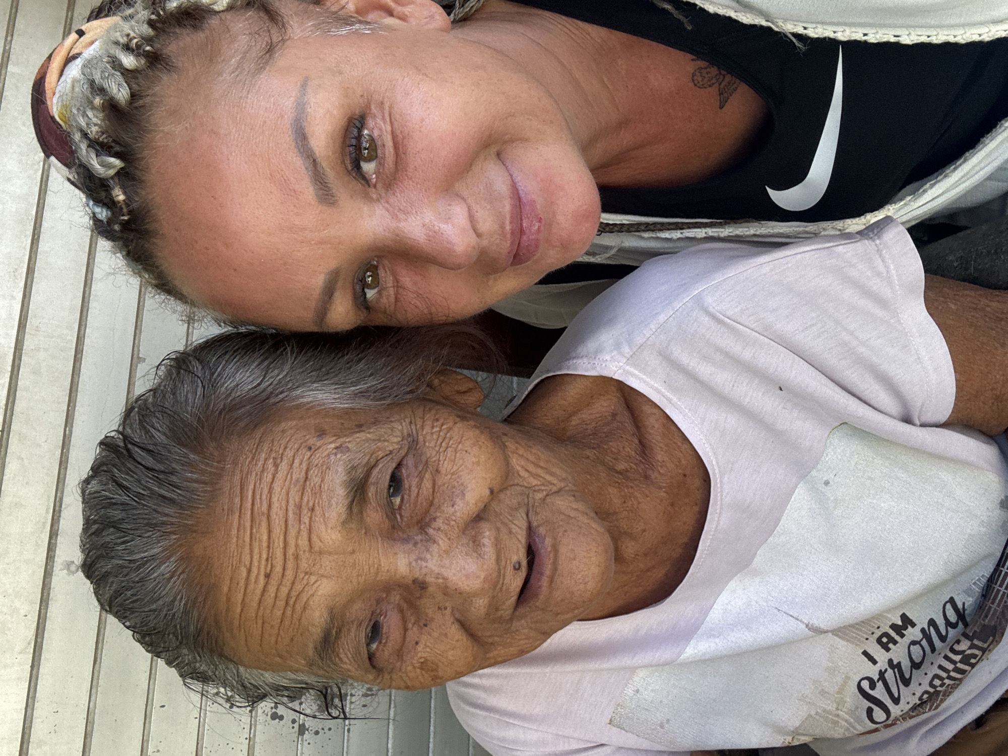 The photograph depicts two individuals in close proximity. The person on the left, who appears younger, has light skin, hazel eyes, and braided hair with a colorful scarf wrapped around their head. They are wearing a black Nike top under a white cardigan, and a small tattoo is visible on their arm. To the right is an older individual with weathered skin, gray hair, and a light pink t-shirt with the words "I AM Strong." The background suggests an outdoor setting in Kuta, Indonesia. The presence of a metal grate-like surface indicates it may be a public area.