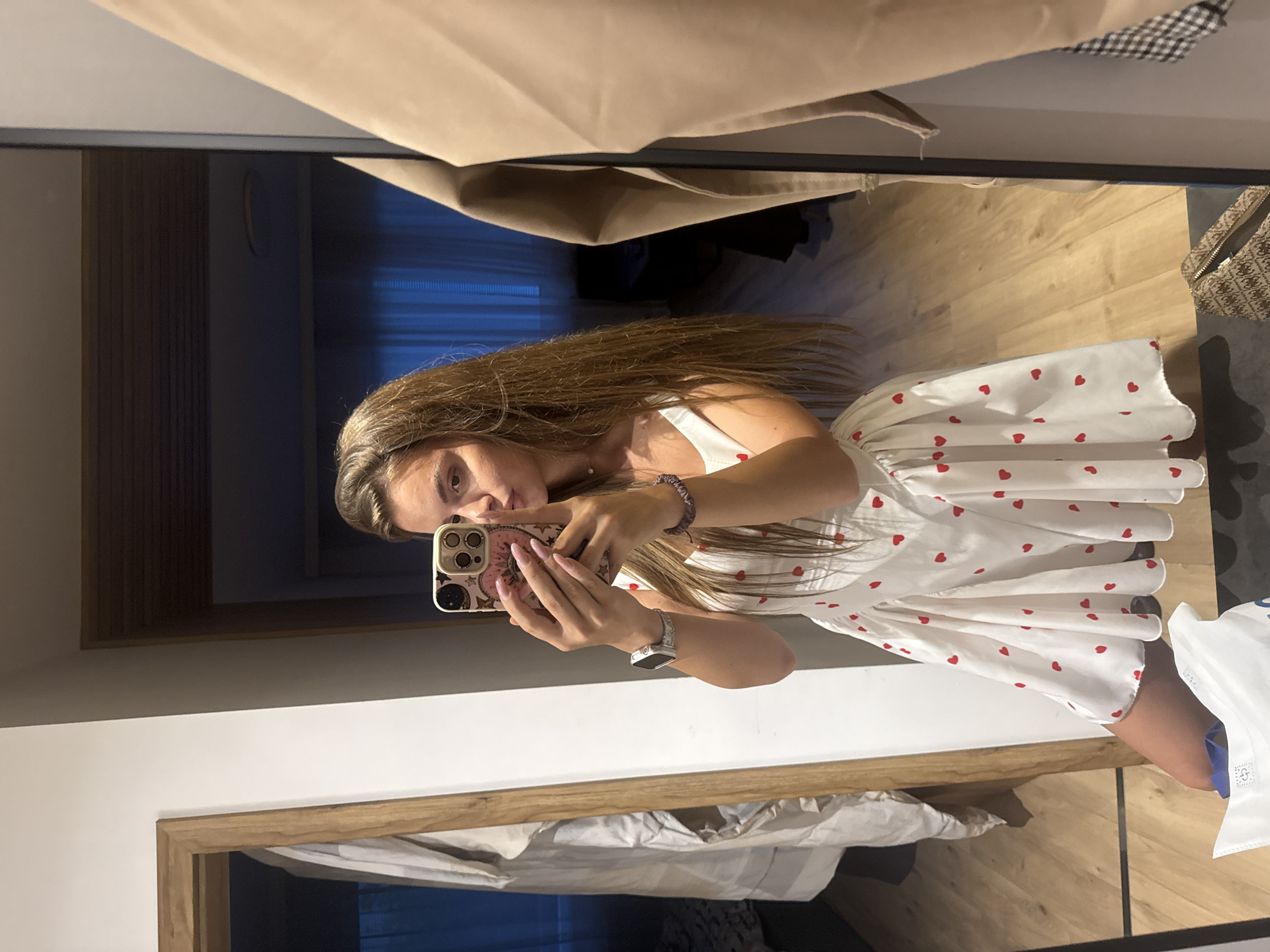 A young woman is captured in a mirror selfie within an indoor setting, identified as Plovdiv, Bulgaria. She stands in a room with light-toned wooden flooring, wearing a white, sleeveless dress adorned with numerous small red heart patterns. Her long, straight brown hair extends down her back and over her shoulders. On her left wrist, she wears a silver-colored smart watch, and on her right, a dark-colored bracelet. Her fingernails are painted pink.
She holds a smartphone with a decorative light-colored case featuring a pink circular design and celestial patterns, positioned in front of the lower half of her face. Her lips are slightly pursed, and her head is tilted. The mirror reflects a segment of a white wall on the left, adjacent to a wooden door frame, which reflects a light-colored garment draped behind it. To the right, a beige garment hangs on a dark rack, with a small portion of a black-and-white checkered pattern visible beneath it. In the background reflection, a darker interior space is visible through an opening, featuring what appears to be dark blue vertical blinds or curtains. On the floor in the foreground, a white shopping bag with blue accents and a brown patterned bag are present.