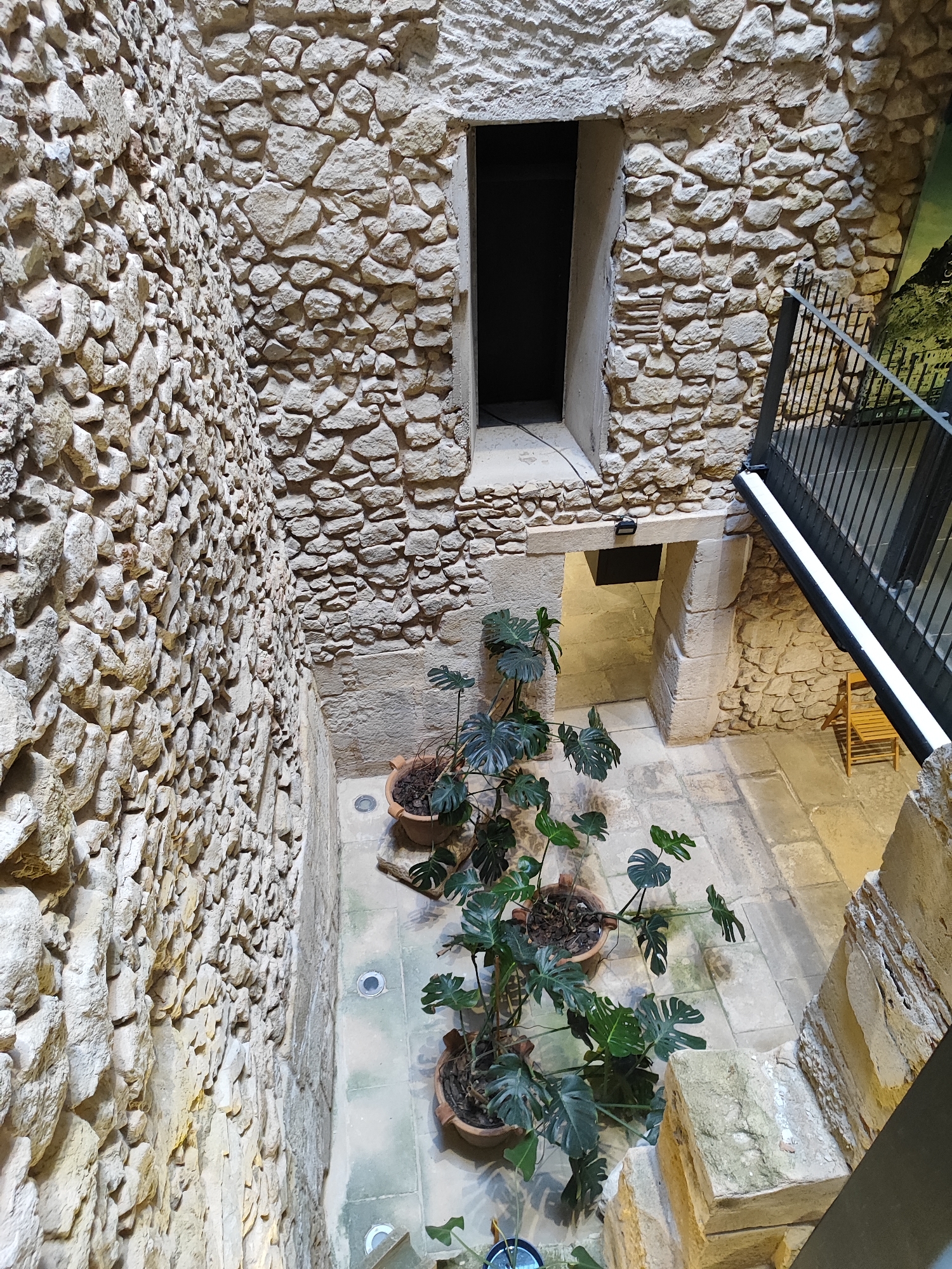 The image depicts an interior courtyard or atrium with rough-hewn stone walls. In the foreground, two large potted plants with broad, split leaves are visible. The plants are situated on the stone-tiled floor. To the right, a black metal railing and a portion of a staircase ascend along the wall. Beyond the plants, a doorway leads into a darker interior space, framed by rough stone. The architecture suggests an old building, possibly a historical site. A single wooden chair is visible through the doorway. The overall impression is of an ancient, well-preserved structure. This image was taken at the Castillo de Santa Bárbara in Alicante, Spain.