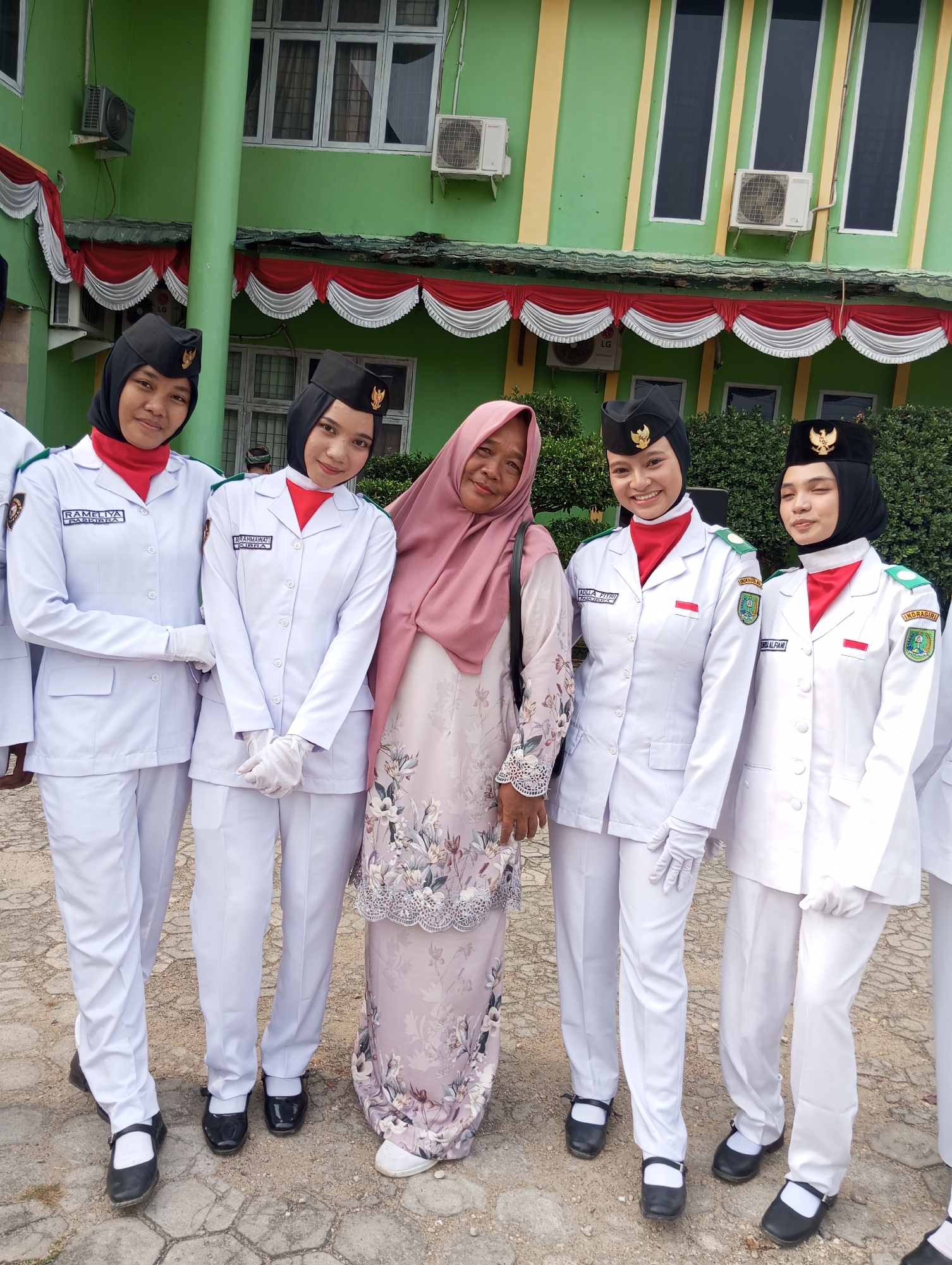 In front of a green building with yellow vertical stripes, five people are posing for a photograph. Four of them are young women in matching white uniforms, black hats with gold emblems, red neck scarves, and white gloves. Their uniforms have name tags and patches on the sleeves. The fifth person is an older woman wearing a pink headscarf and a long, floral-patterned dress. The building has several windows and air conditioning units visible. Red and white bunting decorates the upper facade of the building. The ground is paved with interlocking stones.