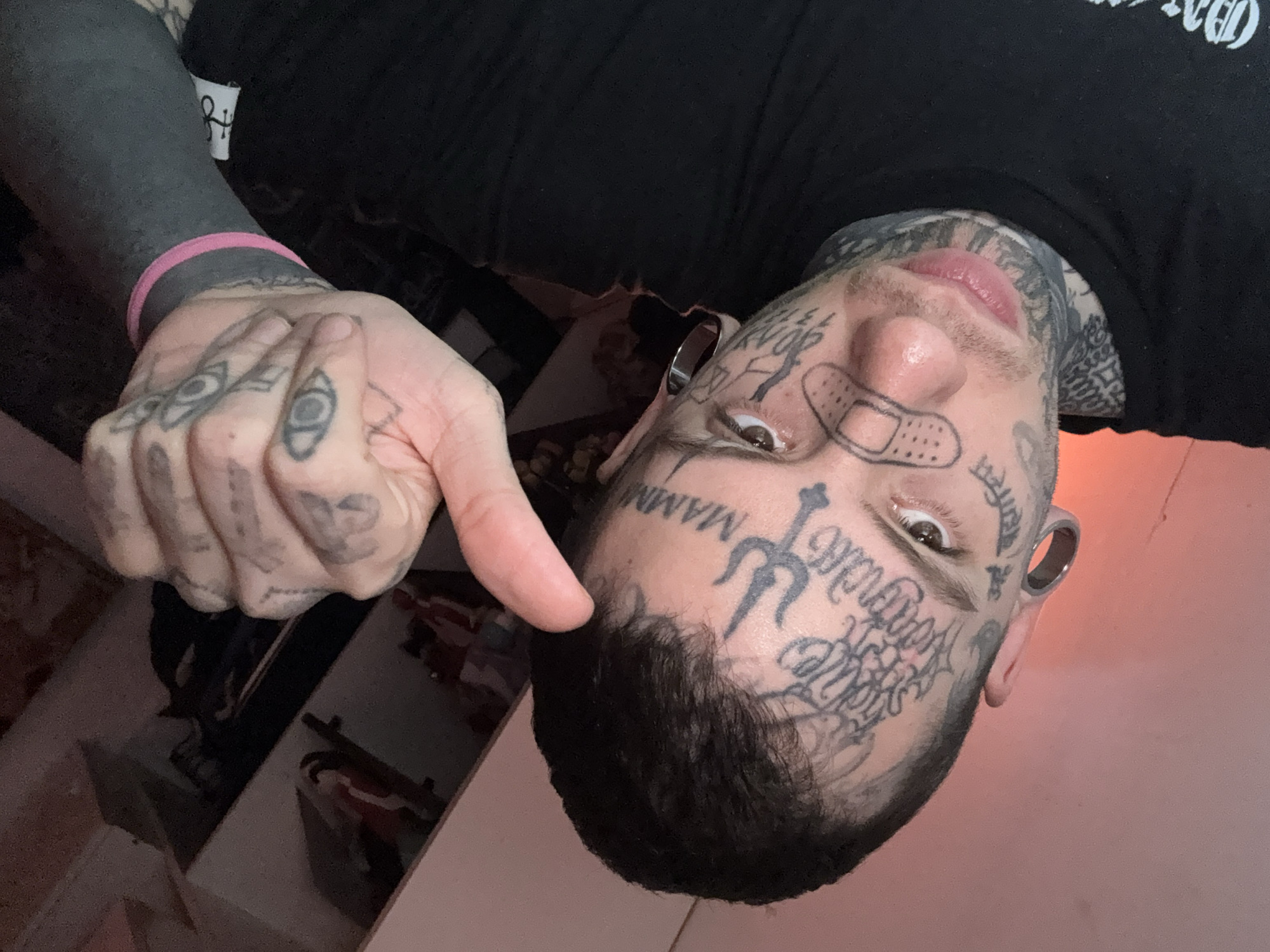A man with extensive tattoos on his face and hands is posing for a selfie. He is wearing a black t-shirt and a pink wristband. His left arm appears to be completely tattooed in black ink. His tattoos include symbols, words, and images, such as a bandage on his nose. He is pointing with his right thumb towards his forehead, which has a trident and the word "MAMMI" tattooed on it. The image is taken indoors, near a shelf unit with various items. The location is not discernible as being within the city of Buenos Aires, Argentina.