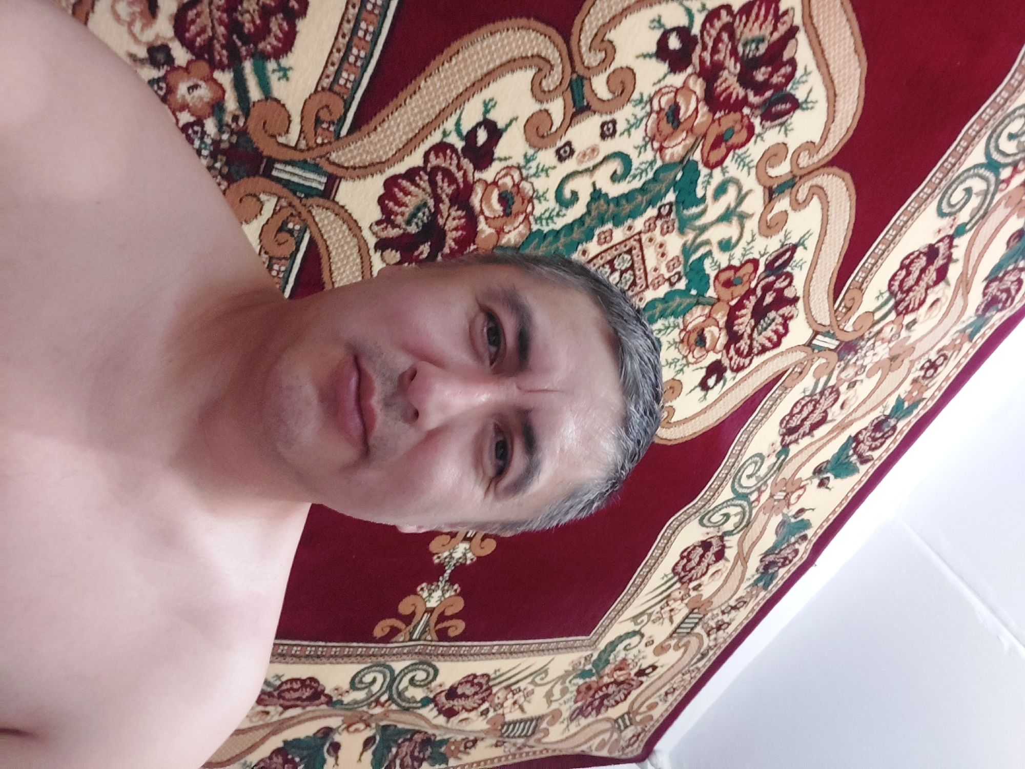 A man with graying hair is pictured from the chest up, looking directly at the camera. He is shirtless, and his face is in sharp focus. The background consists of a large, ornate rug with a maroon border and a cream-colored field featuring floral and scrollwork patterns in shades of red, green, and gold. The lighting suggests an indoor setting, likely a room in a residential building. The image does not provide specific geographical information related to the city of Ması, Kyrgyzstan.