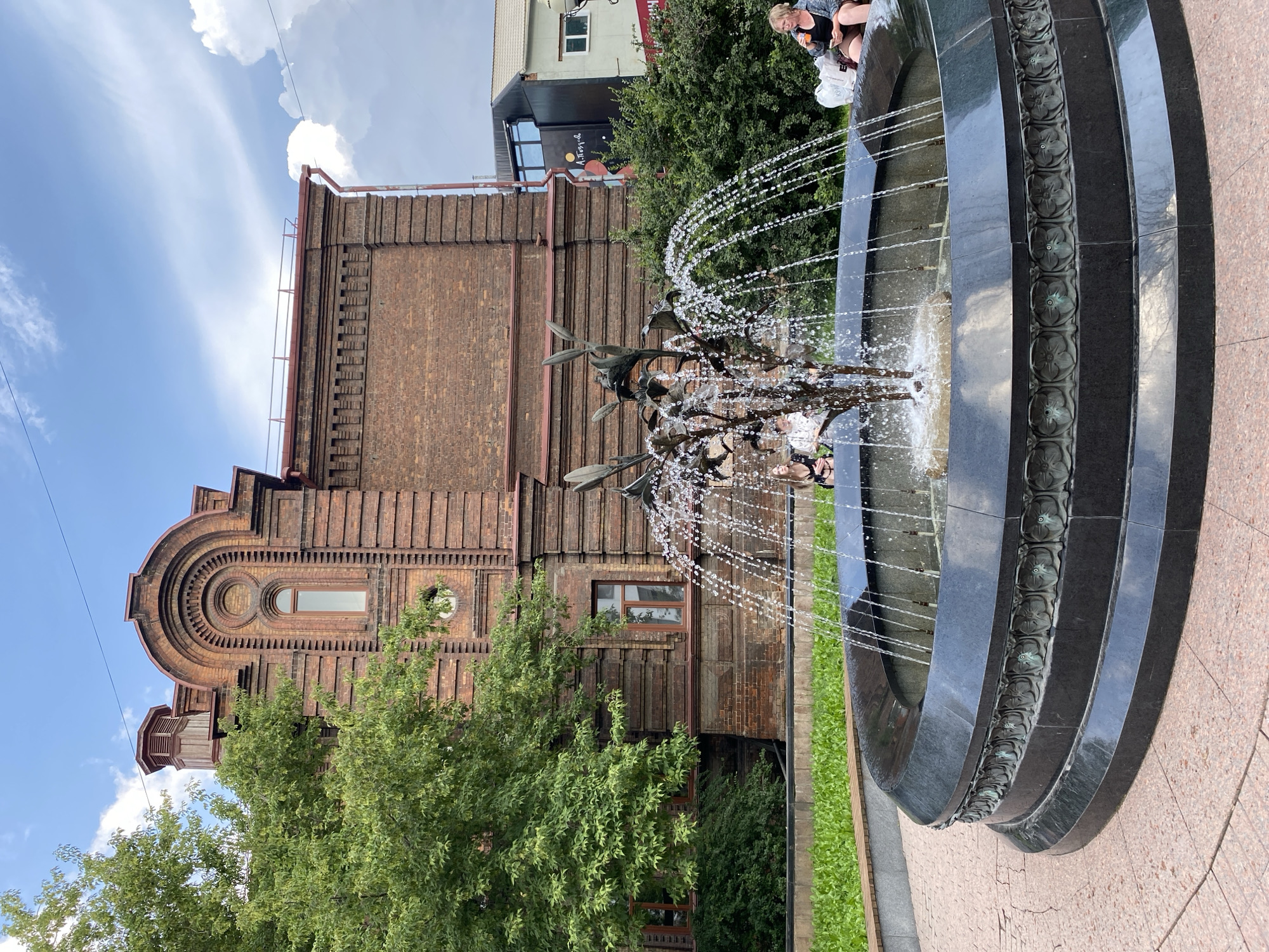 The image shows a fountain in the city of Krasnoyarsk, Russia, located near the Museum of Local Lore. The fountain is centrally located in the foreground, with water spraying upwards from a metallic sculpture resembling branches with leaves. The fountain basin is made of dark stone with ornate carvings along its edge. Behind the fountain, there is a large brick building with distinctive arched windows and architectural details, characteristic of 19th-century industrial architecture. A mature tree with lush green foliage stands to the left of the building. In the background, several people are visible sitting near the fountain and on a bench. One woman on the right is holding a drink and looking at her phone. The sky is cloudy with patches of blue sky. The ground around the fountain is paved with pinkish-red interlocking stones.
