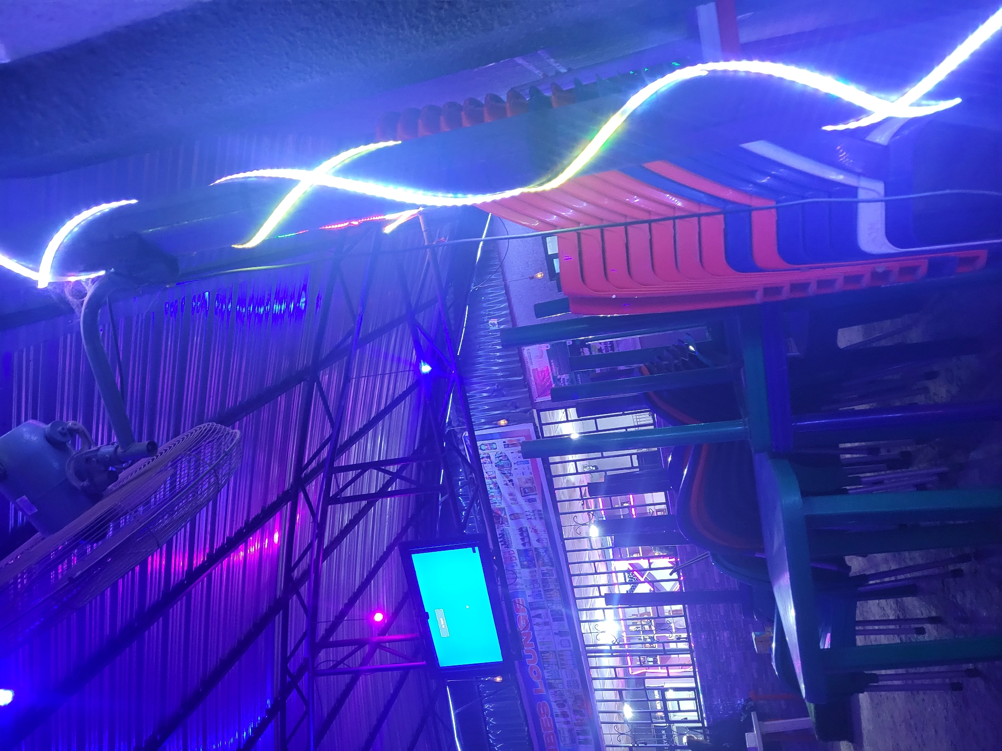 The image depicts a dimly lit outdoor seating area of what appears to be a lounge or restaurant in Madalla, Nigeria. The scene is illuminated by vibrant purple and blue neon lights, creating a nocturnal ambiance. A large industrial fan is mounted on the ceiling, directing air downwards. In the background, a television screen displays a bright turquoise image. Stacks of chairs, primarily orange and blue, are visible, suggesting the area may be closed or preparing for patrons. The ground is covered in sand or gravel. A sign that reads "VIBES LOUNGE" is partially visible above a metal-grated barrier. The overall impression is one of a commercial establishment setting up for evening service or winding down after a busy period.