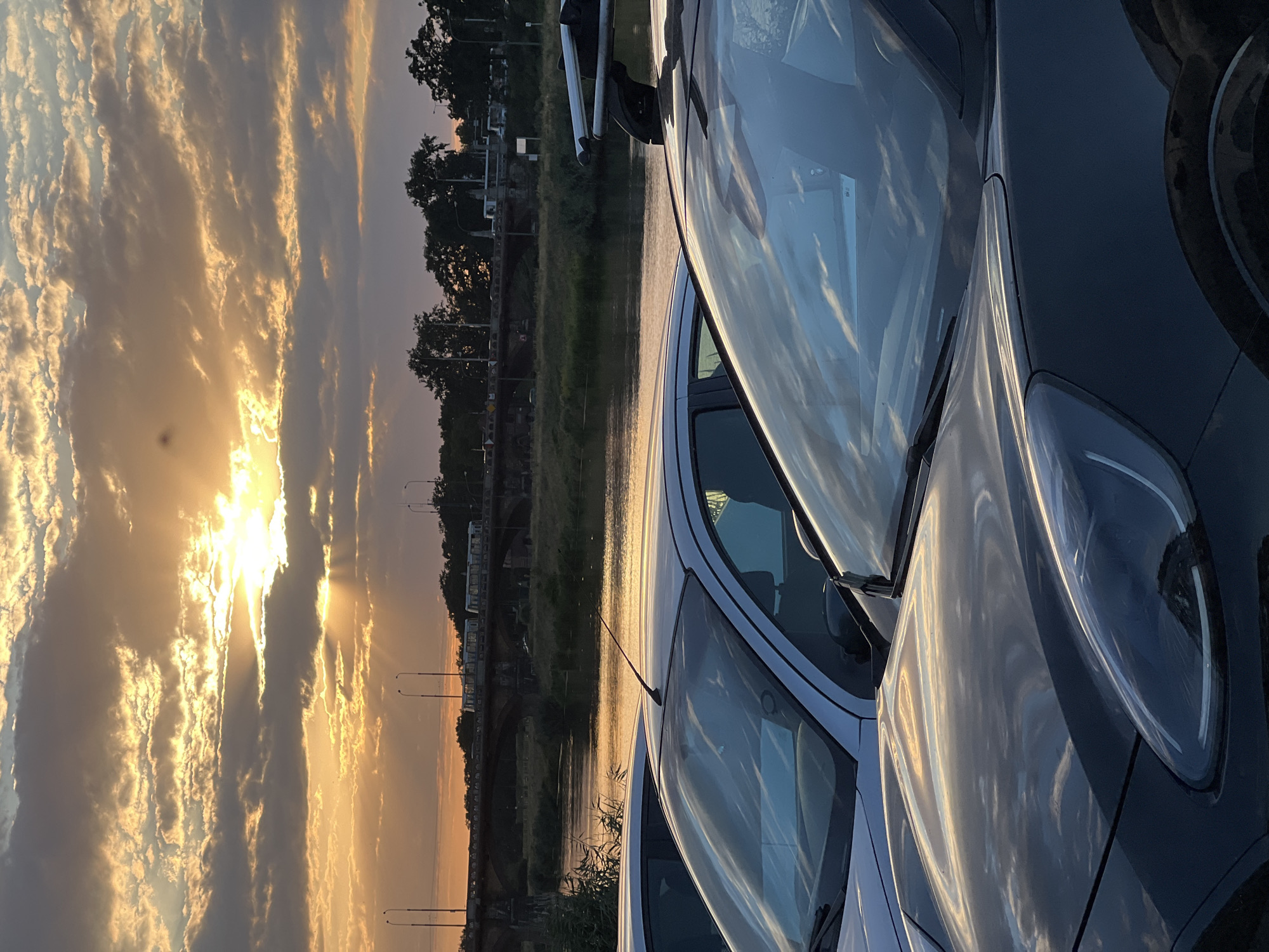The image depicts a sunset scene over the Oder River in Wrocław, Poland. In the foreground, the hoods and windshields of two parked cars are visible, reflecting the golden light of the setting sun and the cloudy sky. The cars are facing towards the viewer. Beyond the cars, the Oder River flows, its surface shimmering with the reflected colors of the sunset. A bridge with traffic, including what appears to be a tram, spans the river in the middle ground. Trees and streetlights line the riverbank. The sky is filled with dramatic clouds, illuminated by the sun which is partially obscured by them, casting rays of light downwards. The overall mood is tranquil and picturesque, typical of a late summer evening.