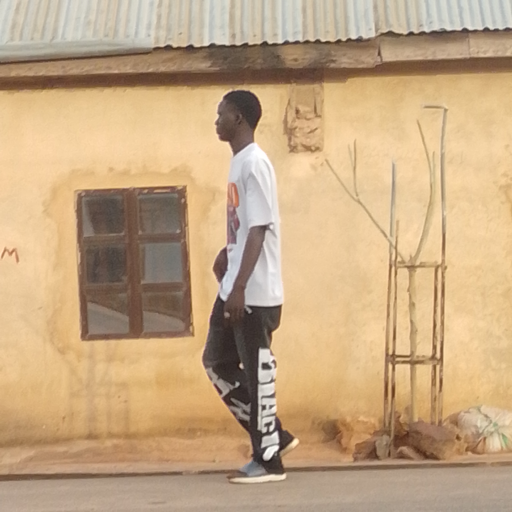 A young Black male is walking in profile to the left, wearing a white t-shirt with an orange and white graphic on the chest, and dark pants with large white lettering down the sides. He is wearing blue and white sneakers. He is walking past a light yellow building with a corrugated metal roof. To the left of the male is a small, dark-framed window with six panes. To the right of the male is a metal structure, possibly a trellis or support for a plant, with some bare branches. There are rocks and a white plastic-wrapped object on the ground to the right of the metal structure. A faint red letter "M" is visible on the building to the left of the window. The scene appears to be a street in Tempure, Nigeria.