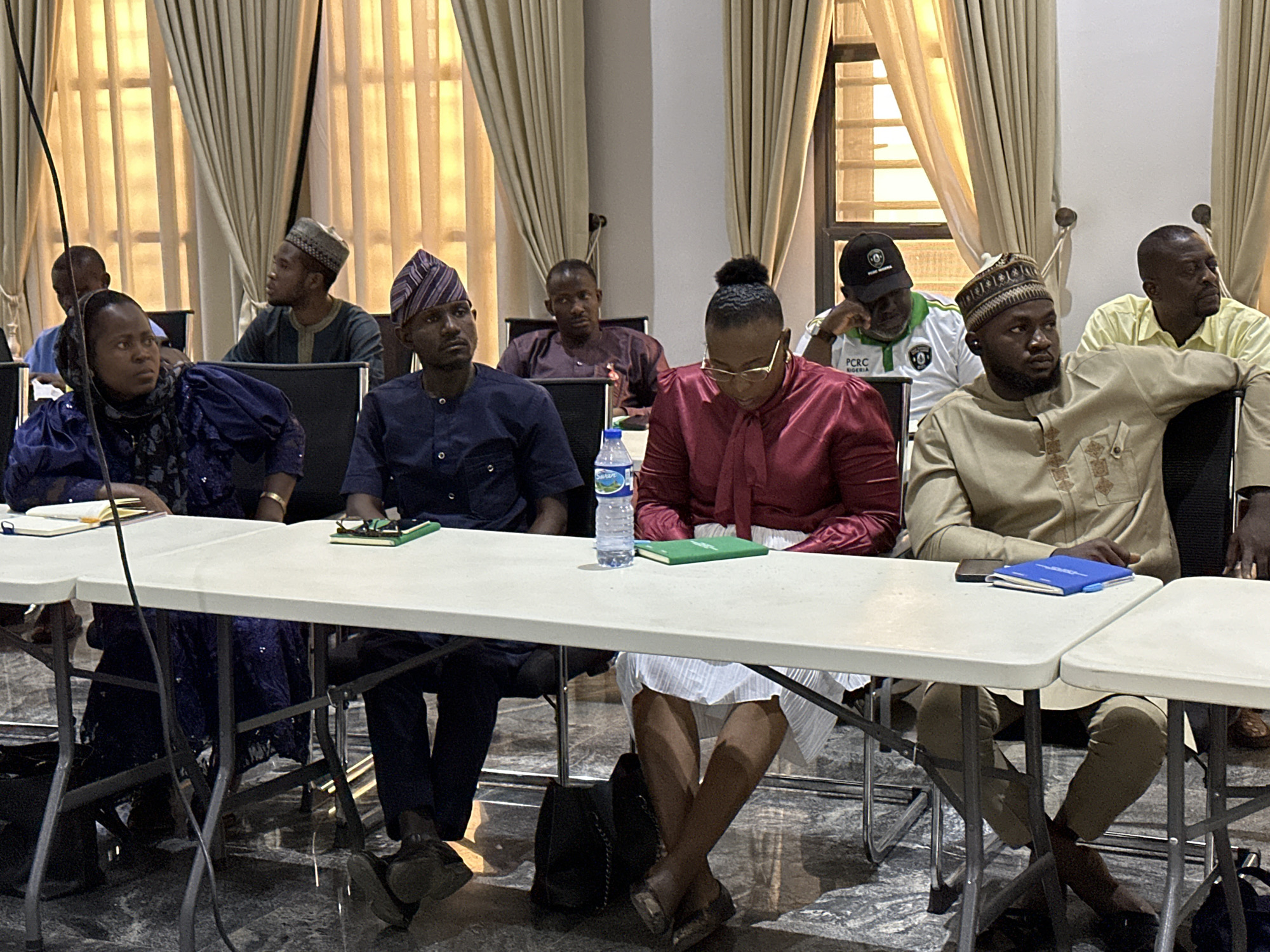 A group of people are sitting at tables in a room with light-colored curtains, possibly for a meeting or conference. The location is in the city of Unknown, Unknown.
There are at least ten individuals visible. They are wearing a variety of clothing, including traditional African attire, such as caps and robes. Some are wearing western-style clothing like button-down shirts. Several people have notepads or notebooks on the tables in front of them.
The room has neutral colored walls and the tables are plain white. A water bottle sits on the table near the center. The scene appears to be indoors with natural light filtering through the windows.
