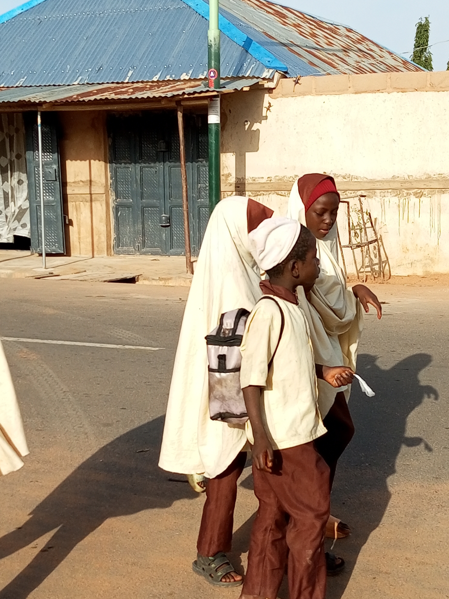 In Tempure, Nigeria, two children are walking on a paved road. The child in the foreground, on the right, is wearing a cream-colored tunic and brown trousers. They have a white cloth in their right hand and are looking to their left. The child behind them, on the left, is wearing a long cream-colored garment that covers most of their body, and a white head covering. They have a bag with a black and grey patterned strap hanging from their left shoulder. The children appear to be walking together.
In the background, there is a building with a corrugated metal roof that is partially rusted. The building has large, dark-colored doors and some white curtains visible. A green pole stands to the right of the children. The ground on the side of the road is a mix of dirt and pavement. Shadows of the children are cast on the road, indicating it is a sunny day.
