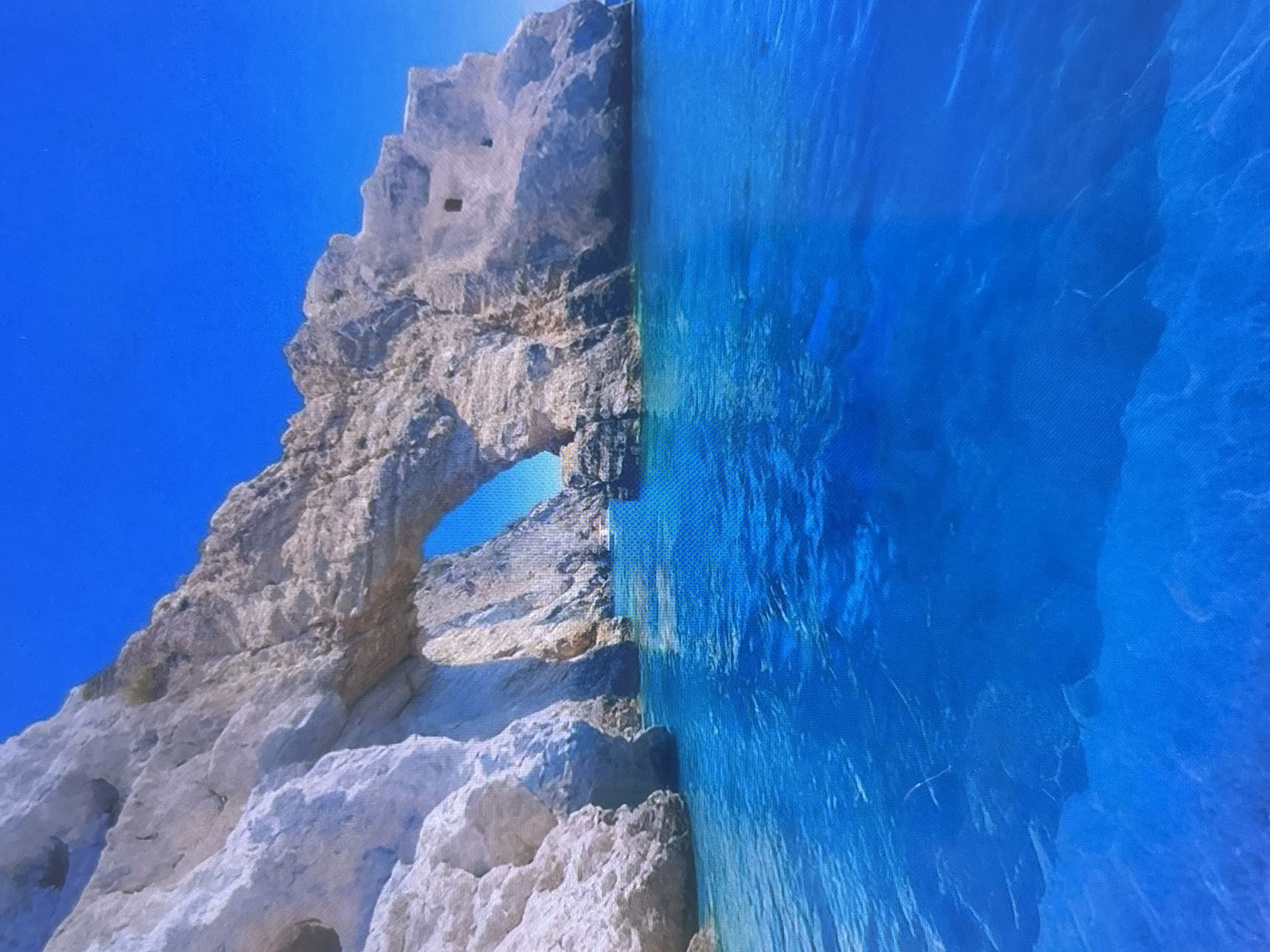 The image, oriented vertically, depicts a stunning natural coastal landscape under a bright, clear sky. On the left, a large, rugged cliff face, composed of light-colored, weathered rock with varying shades of gray and brown, dominates the scene. The cliff features intricate textures, numerous crevices, and several small, dark, cave-like openings. A prominent natural rock arch is visible in the lower part of the cliff, through which the brilliant blue sky or distant water can be seen.
To the right, an expansive body of water, transitioning from a vibrant turquoise near the shore to a deeper blue further out, meets the rocky cliff. The water appears exceptionally clear, with subtle ripples and reflections on its surface. The overall impression is one of calm, pristine marine conditions.
The sky above the cliff and across the top of the frame is a pure, cloudless azure, suggesting a bright and sunny daytime setting. There are no people or man-made objects visible, highlighting the untouched natural beauty of the location. The image exhibits a subtle grid pattern, indicating it is likely a photograph taken of a digital screen.