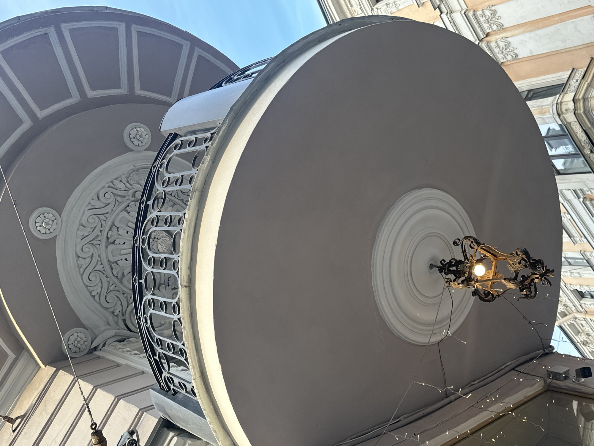 This is a low-angle, outdoor photograph of an ornate balcony on a building in Saint Petersburg, Russia. The balcony is curved and has a light pink soffit. A black metal railing with oval openings surrounds the top of the balcony. Above the balcony, architectural details include decorative moldings, floral motifs, and a recessed ceiling with square patterns. A vintage-style wrought-iron lantern with a lit bulb hangs from the center of the soffit. String lights are draped along the underside of the balcony's edge. The background shows parts of adjacent buildings with classical architectural features, including cream-colored facades with orange accents, window frames, and decorative cornices. The sky is clear and blue. There are no people visible in the frame.