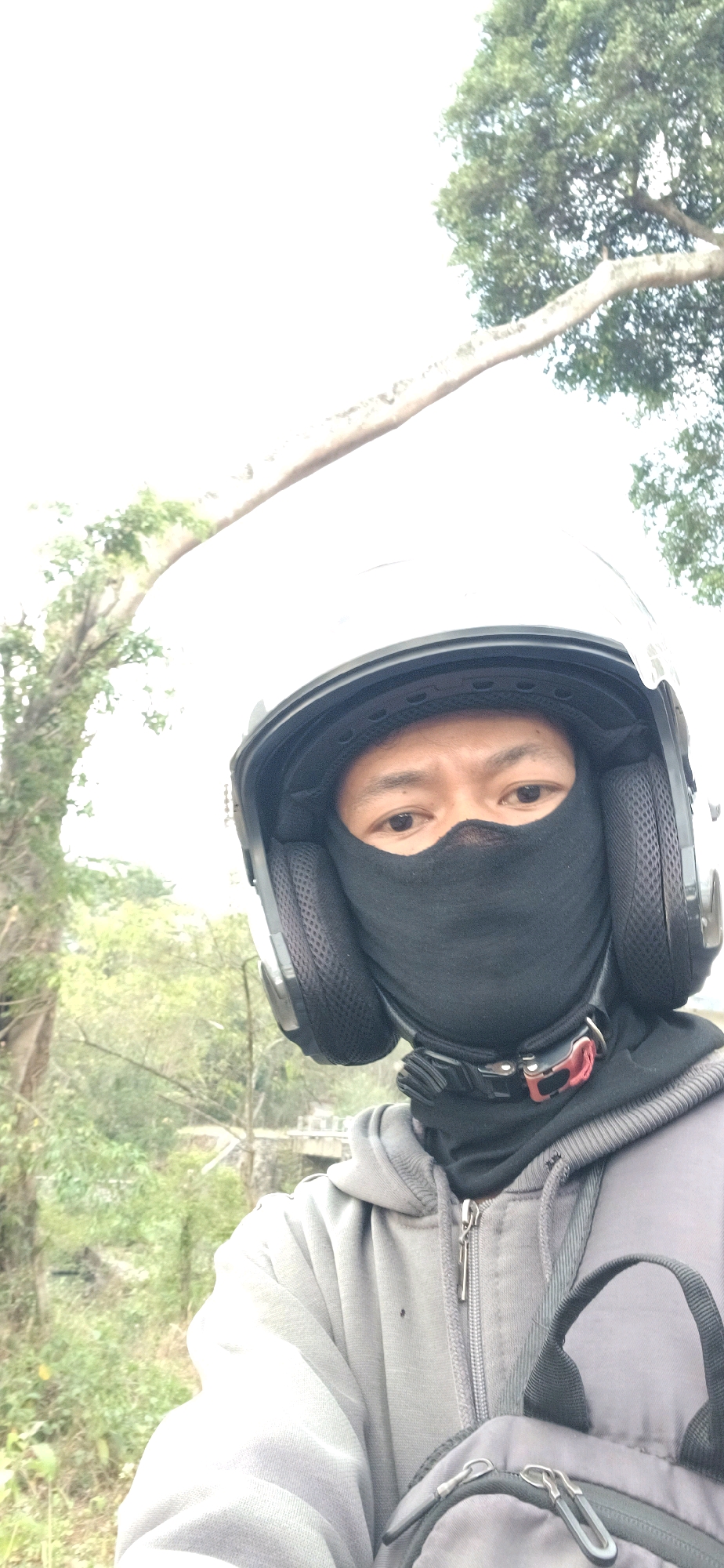 A person wearing a white helmet, black balaclava, and a grey hoodie is taking a selfie outdoors. The background shows trees and what appears to be a bridge in the distance. The person's eyes are visible through the balaclava. The scene is likely in Tengahan, Indonesia, based on the provided context.
