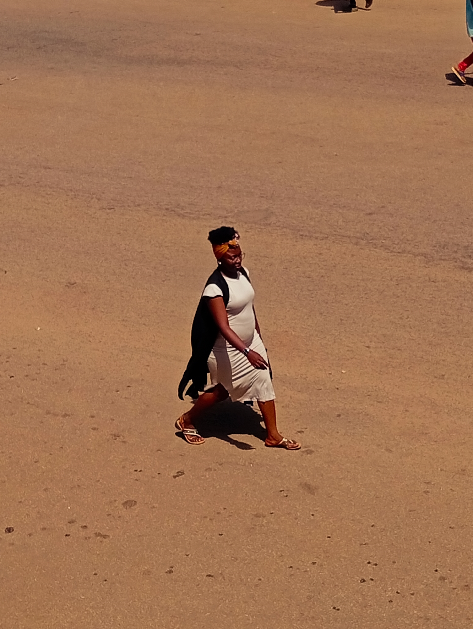 A woman with a head wrap and glasses is walking across a dusty, light brown asphalt surface in Bukuru, Nigeria. She is wearing a white dress and a dark vest. The woman is in motion, with her left leg extended forward. The sun is casting a shadow to her left. There are a few dark spots scattered across the surface. In the background, there are faint shadows of other people and their legs, indicating a public area.