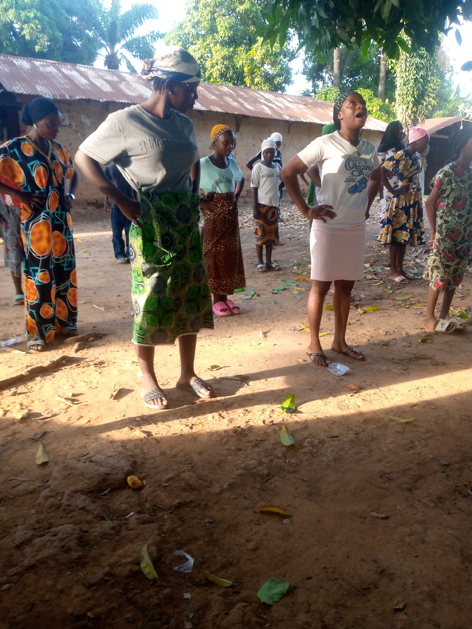 A group of at least 10 individuals, predominantly female, stand on a dirt ground with scattered leaves. The location is an outdoor area possibly in the city of Unknown, Unknown. The people appear to be in casual clothing, with some wearing patterned fabrics wrapped around their waists. The person in the middle is looking up with her mouth open. The background includes trees, foliage, and a building with a corrugated metal roof.