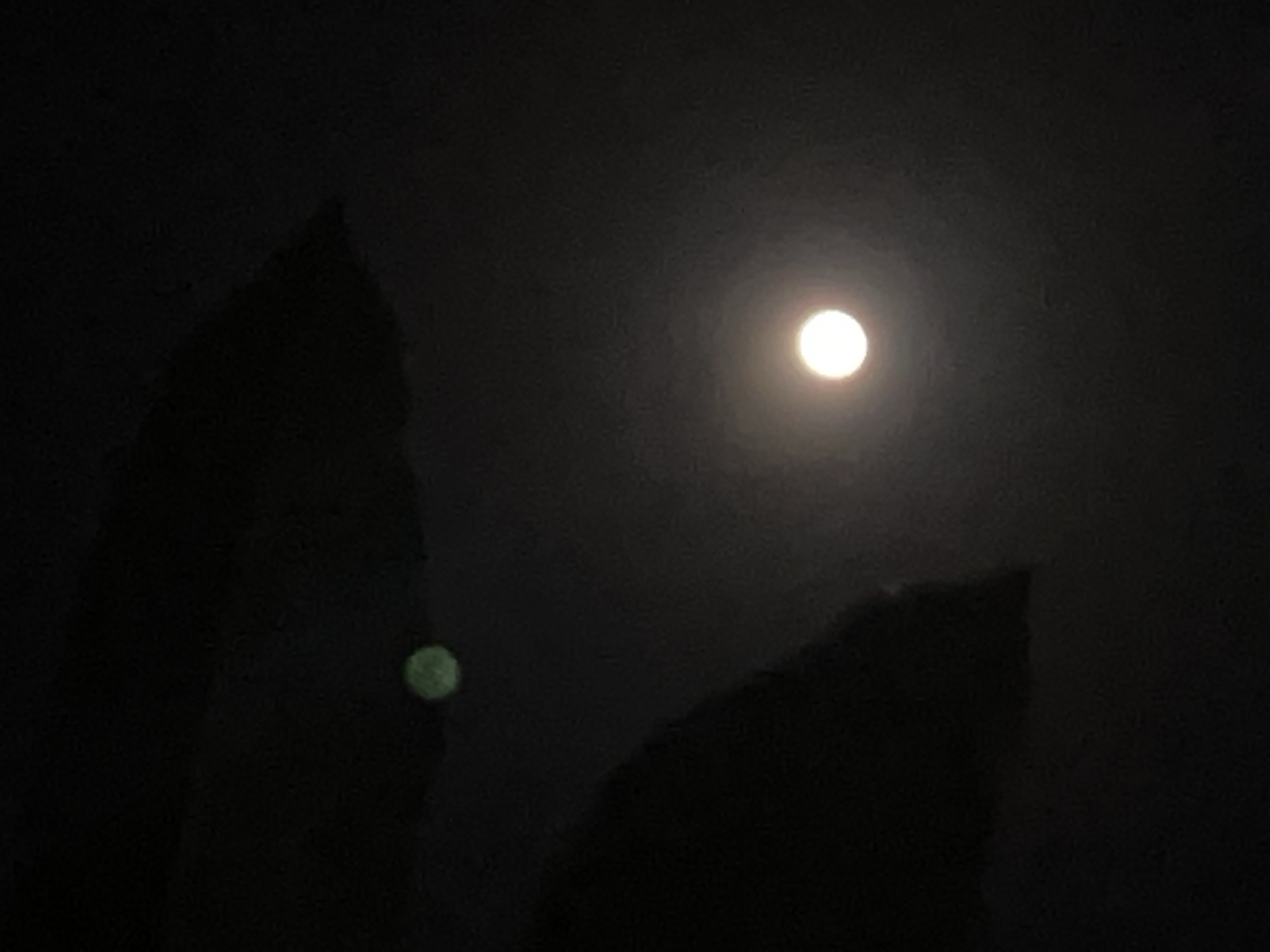 The image shows a dark night scene, likely taken in Canggu, Indonesia. A bright moon is visible in the upper center of the frame. Silhouetted objects, possibly trees or buildings, occupy the lower portions of the image. A small, indistinct green light is visible on the left side, seemingly on one of the objects.