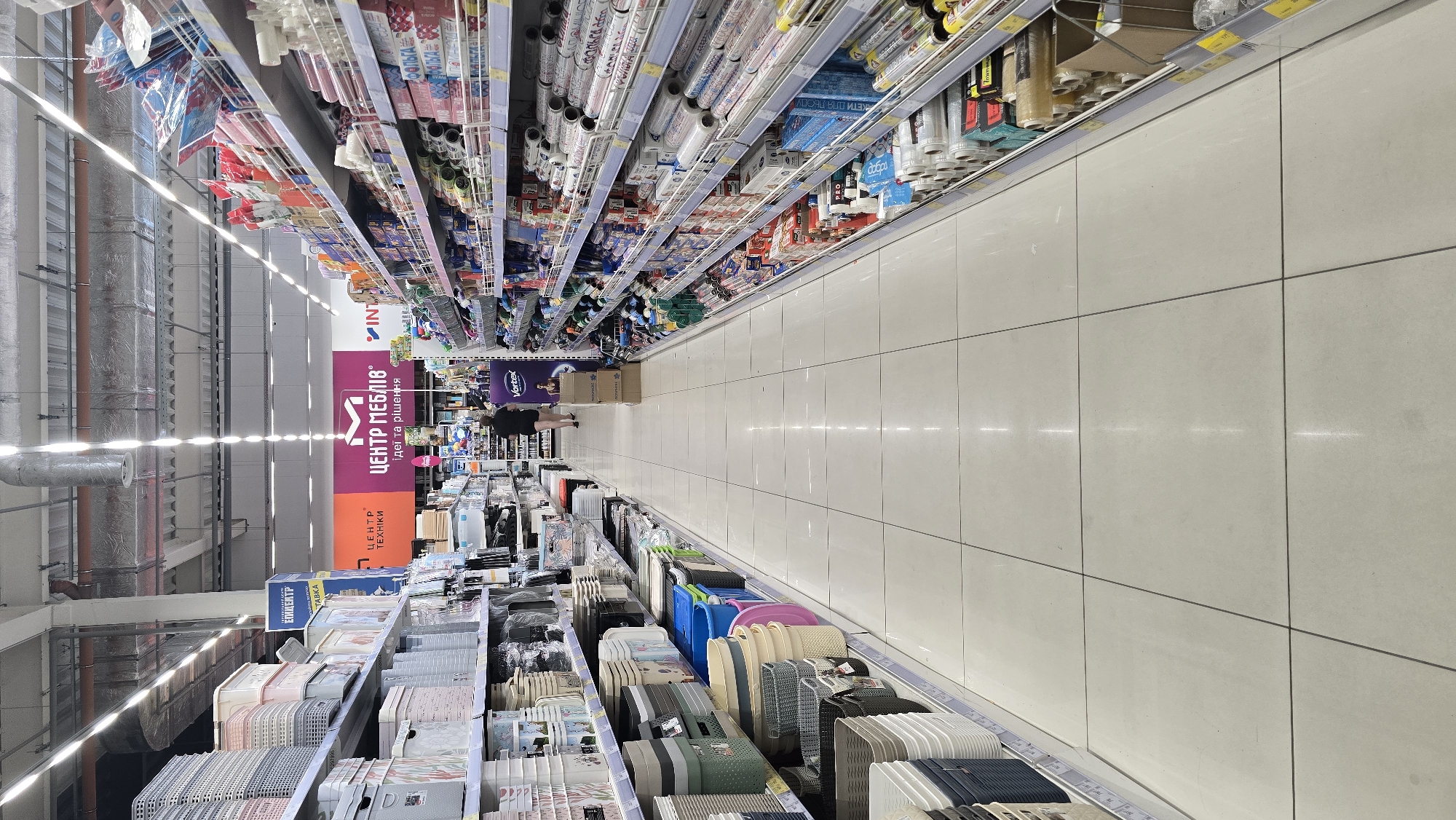 The image captures an interior view of a brightly lit supermarket aisle in Zhytomyr, Ukraine, featuring extensive shelving stocked with various household goods. The perspective is from the aisle, looking down its length, with shelves lining both sides. The left side displays an array of plastic storage containers in various sizes and colors, including white, light grey, and some with decorative patterns. These are neatly stacked on multiple tiers. The right side of the aisle is dominated by shelves filled with rolls of wrapping paper, foil, and other similar items, many of which are packaged in colorful plastic or cardboard. Prominent signage is visible overhead, with large letters spelling out "ЦЕНТР МЕБЛІВ" (Furniture Center) and smaller text indicating "ідеї та рішення" (ideas and solutions). A person, dressed in dark clothing, is walking away from the camera down the aisle on the right side, appearing to be a shopper. The floor is tiled with large, pale grey squares that reflect the overhead lighting. The overall environment suggests a retail setting designed for broad consumer goods.