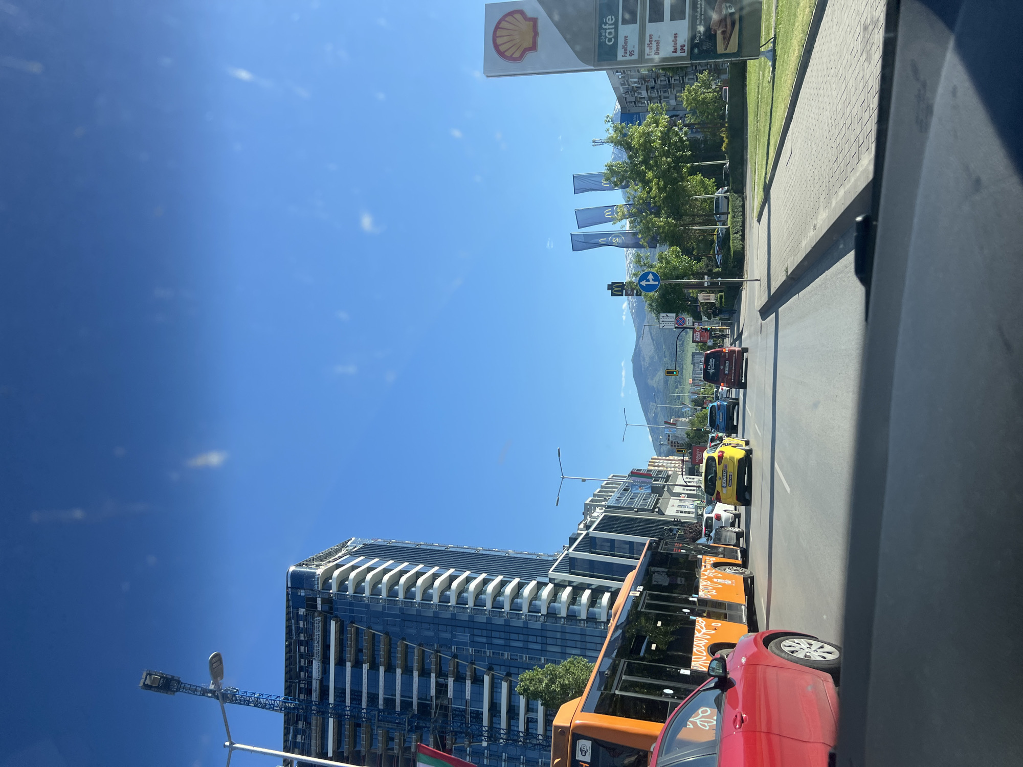 The image shows a street scene in Sofia, Bulgaria, likely taken from inside a vehicle. The view is along a road with a Shell gas station visible on the right side. A tall, modern building is under construction on the left. There is moderate traffic on the road, including a yellow taxi, an orange bus, and a red car closer to the camera. A mountain range is visible in the background, suggesting the proximity to the Vitosha Mountain. There are various street signs, including directional signs and possibly advertisements. The sky is clear and blue. Flags with the McDonald's logo can be seen in the distance, indicating the location of a McDonald's restaurant.