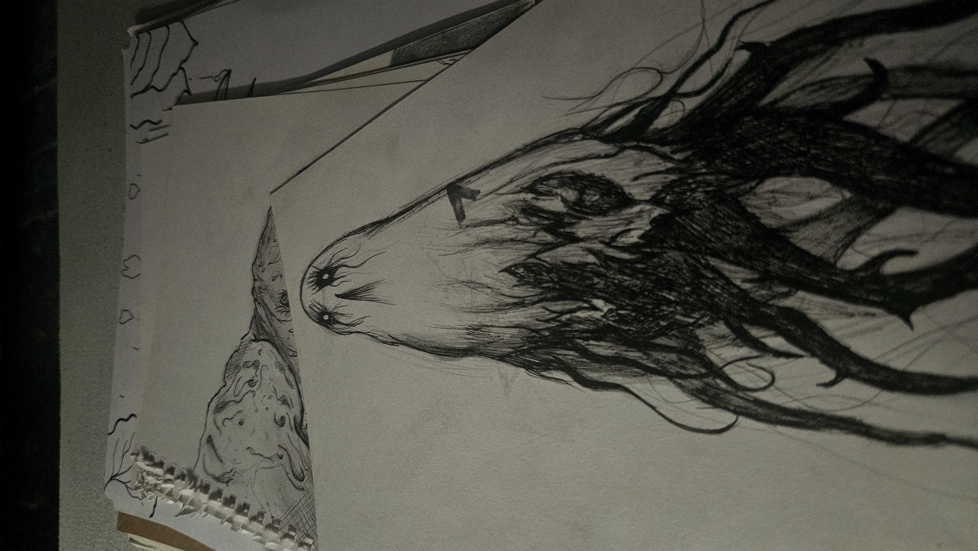 The image is a close-up, high-angle shot of a pencil sketch on a piece of paper. The sketch depicts a dark, abstract, humanoid figure with flowing, wispy hair or tendrils and hollow, glowing eyes. The figure appears to be emerging from a dark, textured base, possibly representing roots or shadows. In the background, another sketch is visible, showing a landscape with swirling clouds or mist. The paper is laid on a surface that appears to be a table or desk. The lighting is dim, creating shadows and highlighting the texture of the sketch. The composition suggests an artistic process, with the sketches appearing to be works in progress.