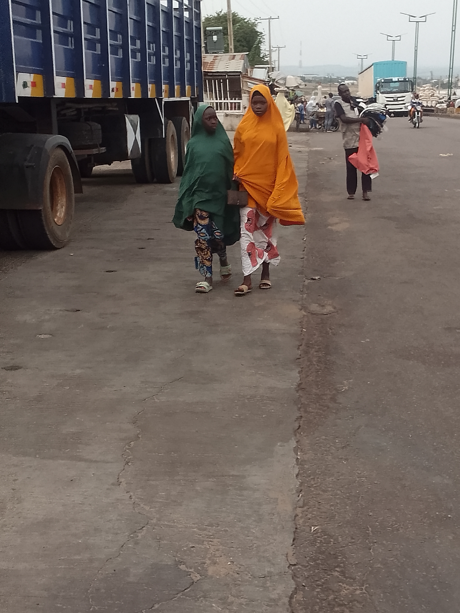 Two young girls, one in a green hijab and the other in an orange hijab, walk side-by-side on a paved road. The girl in green wears a patterned skirt and sandals, while the girl in orange wears a white skirt with red floral patterns and sandals. Behind them, a man carries a bundle of clothes and a pink bag. In the background, a large blue truck is parked, and other vehicles, including a white truck and a motorcycle, are visible on the road. Small stalls or shops line the side of the road, and utility poles are present. The scene appears to be set in a marketplace or a busy roadside area. The lighting suggests it could be daytime, possibly late afternoon or early morning, with overcast conditions. The overall atmosphere seems to be one of daily activity in a bustling environment.