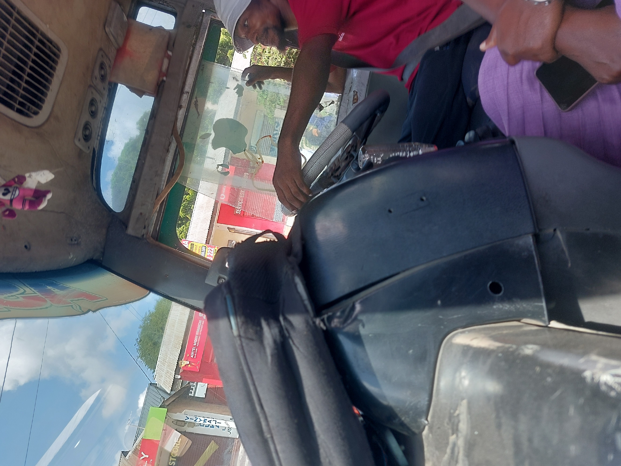 The image captures the interior of a vehicle, possibly a bus or van, with a man in a red shirt driving. He is wearing a white hat and has his hand on the steering wheel. A passenger in a purple shirt is seated next to him, holding a phone.
Through the windshield, the street scene is visible, showing commercial buildings with signs in Swahili. "Airtel Money Branch" is visible in red, and "STOORE" and "VINYWAJI" on storefronts. The general location appears to be Dar es-Salaam, Tanzania based on the language and style of signage.