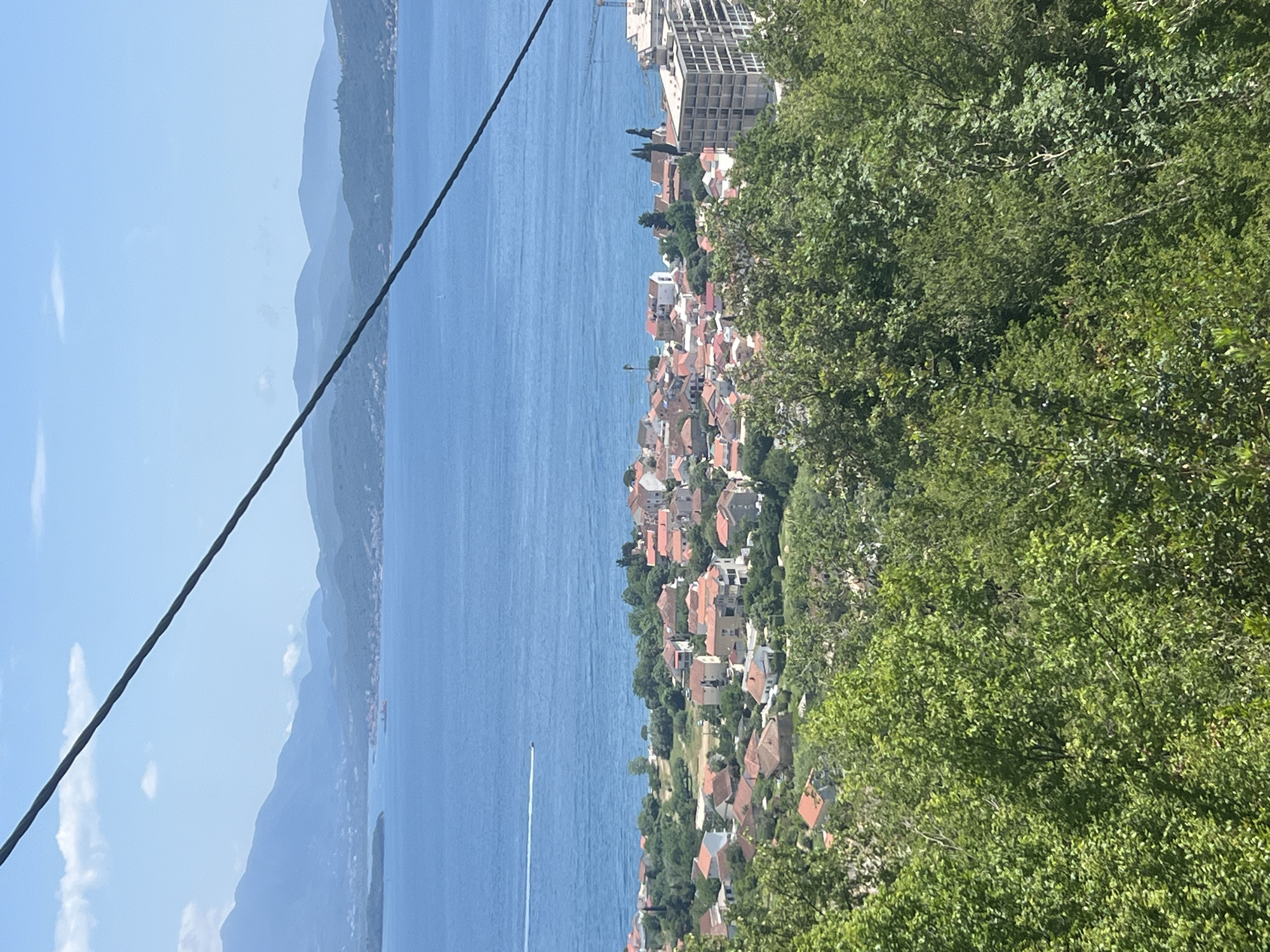 The image shows a coastal landscape in Baošići, Montenegro. The scene is captured from an elevated viewpoint, likely a hillside, overlooking the Bay of Kotor. The body of water is light blue and stretches into the distance, bordered by mountains with sparse vegetation. The mountains fade into the distance with a blue haze. A cable runs diagonally across the left side of the frame. A coastal town with red-tiled roofs lines the shore. A modern, multi-story building is visible among the town's structures. Dense greenery, including trees and bushes, fills the right side of the frame. A small boat leaves a white wake in the sea. The sky is light blue with minimal cloud cover.