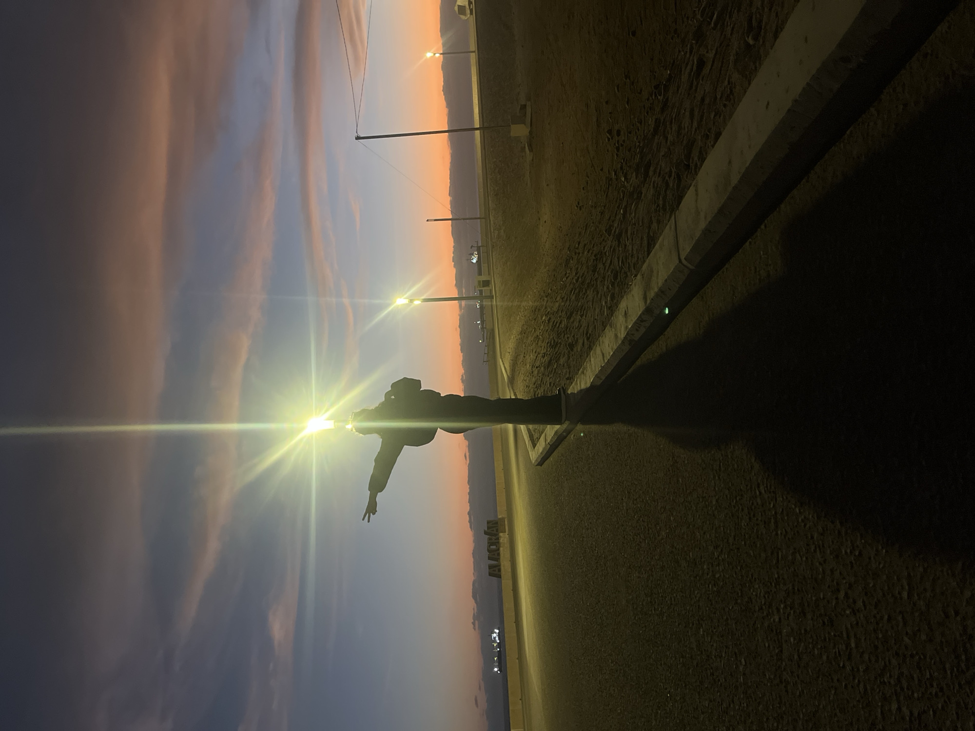 The image depicts a landscape in Arica, Chile, at dusk or dawn. The sky features a gradient of colors from dark blue/purple to orange, with some cloud formations. A person, seen in silhouette, stands on a road next to a concrete barrier. The road is illuminated by street lights, and their light creates a bright glare in the image. Several lamp posts are visible along the road. The name "ARICA" in large letters is seen from the side of the road. In the distance, faint lights suggest a town or city.