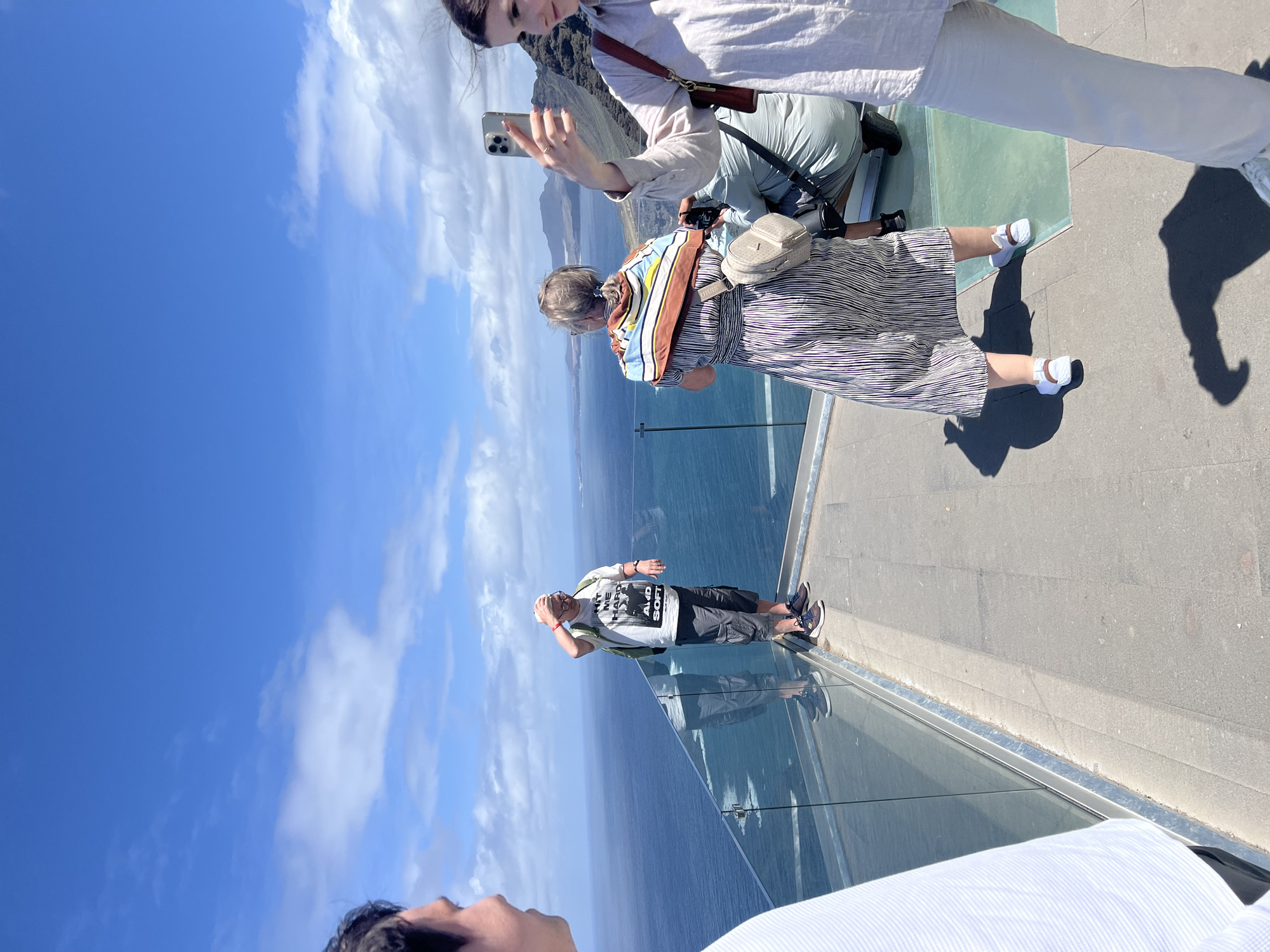 The image shows a group of people at the Barranco de Guayedra viewpoint in Spain. Two women are taking photos; one is using a phone, while the other is taking a photo of the scenery. A man stands near the edge of the viewing platform with the ocean and sky behind him. The viewpoint has a glass floor and clear railings, providing unobstructed views. The day is sunny, with a blue sky and some clouds. The environment consists of the ocean, cliffs, and the sky.