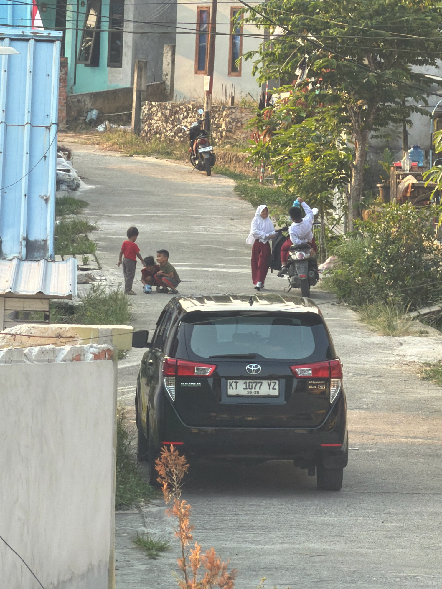 In a residential area of Balikpapan, Indonesia, a black Toyota Innova is parked on an uphill road. Three young boys are gathered on the asphalt, one standing and wearing a red shirt, while two are crouched down, one appearing to be interacting with a toy. Further up the road, two individuals, a child wearing a white headscarf and a dark outfit, and another person in a white shirt, are near a parked motorcycle. The child in the white headscarf is walking, while the other individual is seated on the motorcycle. Buildings with various architectural styles line the sides of the road, some appearing to be under construction or in disrepair, with vegetation growing around them. The scene is illuminated by sunlight, casting shadows and suggesting it is either early morning or late afternoon.