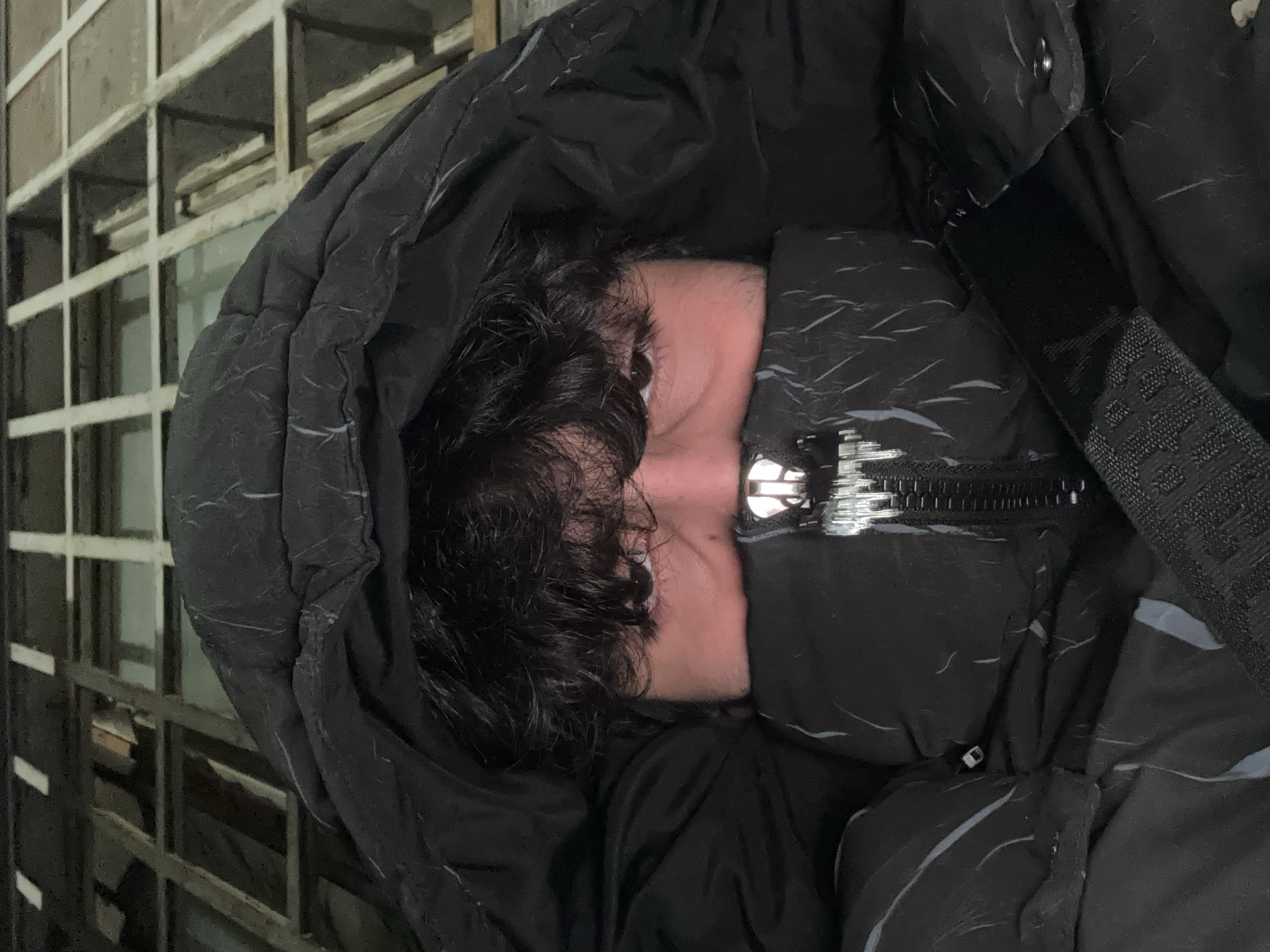 A person is seen in the image wearing a black, hooded puffer jacket, with the hood pulled up, obscuring most of their face except for the eyes and some of their dark, curly hair. The jacket is zipped up. A shoulder strap is visible on the right side of the image. The background appears to show a metallic or barred structure. The image was likely taken in Yerevan, Armenia.