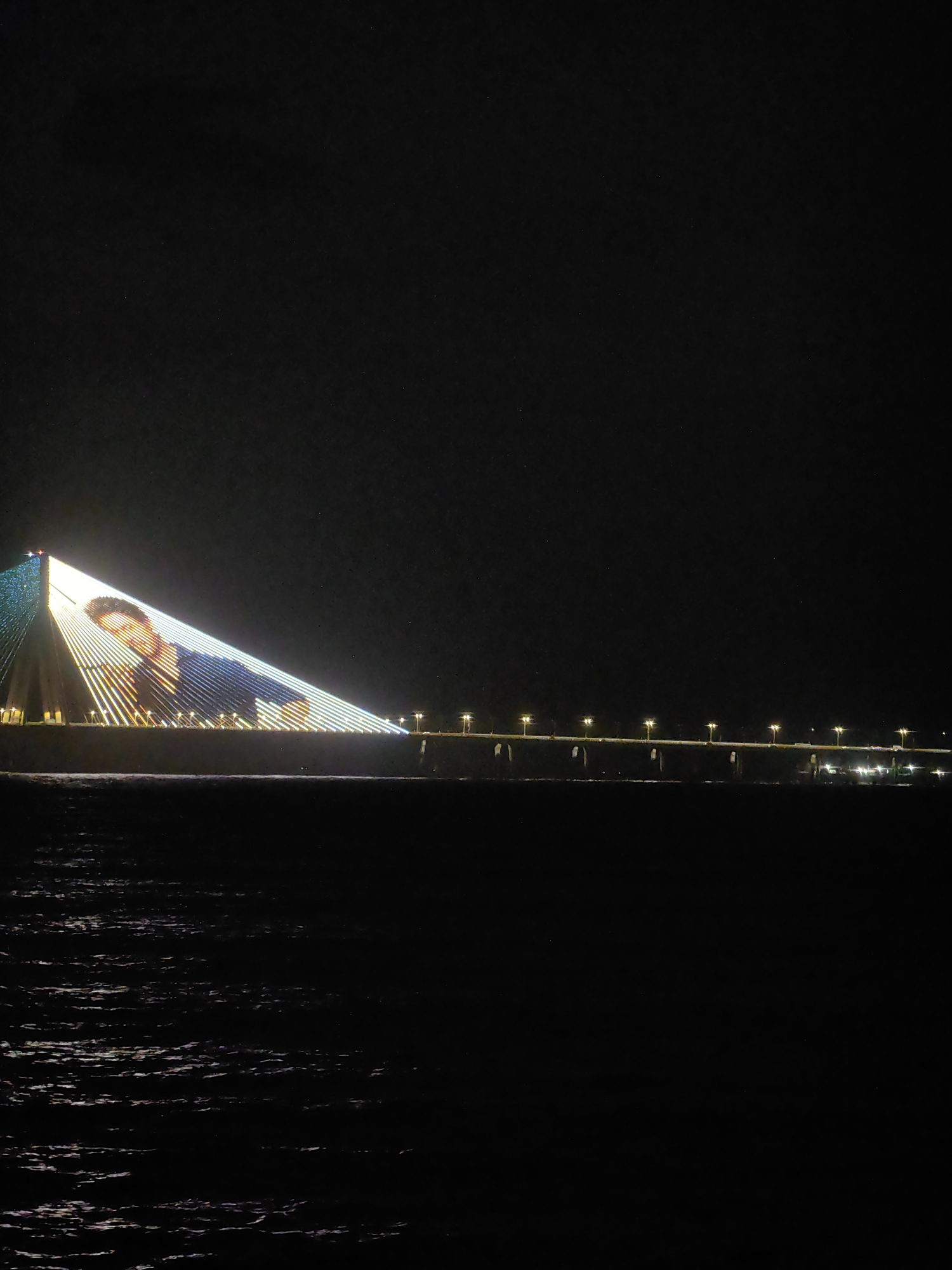 The image shows a nighttime view of the Bandra-Worli Sea Link in Mumbai, India. The left pylon of the bridge is illuminated with a large projection of a man's face. The bridge itself is lit with strings of lights that form a diagonal pattern leading up to the pylon. The dark water of the Arabian Sea is visible in the foreground, reflecting the lights of the bridge and the city in the distance. The sky is black, indicating it is nighttime.