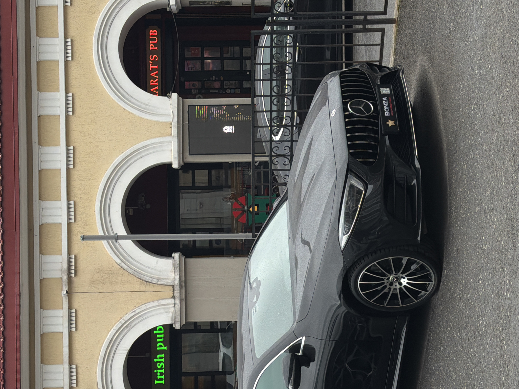 A black Mercedes-Benz sedan is parked on a wet asphalt street in front of a building with arched entrances. Above the entrances, neon signs read "IRISH PUB" in green and "ARAT'S PUB" in red. The building appears to be a commercial establishment, possibly a pub or bar, with dark windows. A black metal fence separates the street from the building's entrance area. A person is visible through the glass door on the right side of the "ARAT'S PUB" entrance, wearing a red garment. The sky is overcast, suggesting a cloudy or rainy day. The image appears to be taken in Ufa, Russia, as indicated by the license plate on the car which is typical for that region.