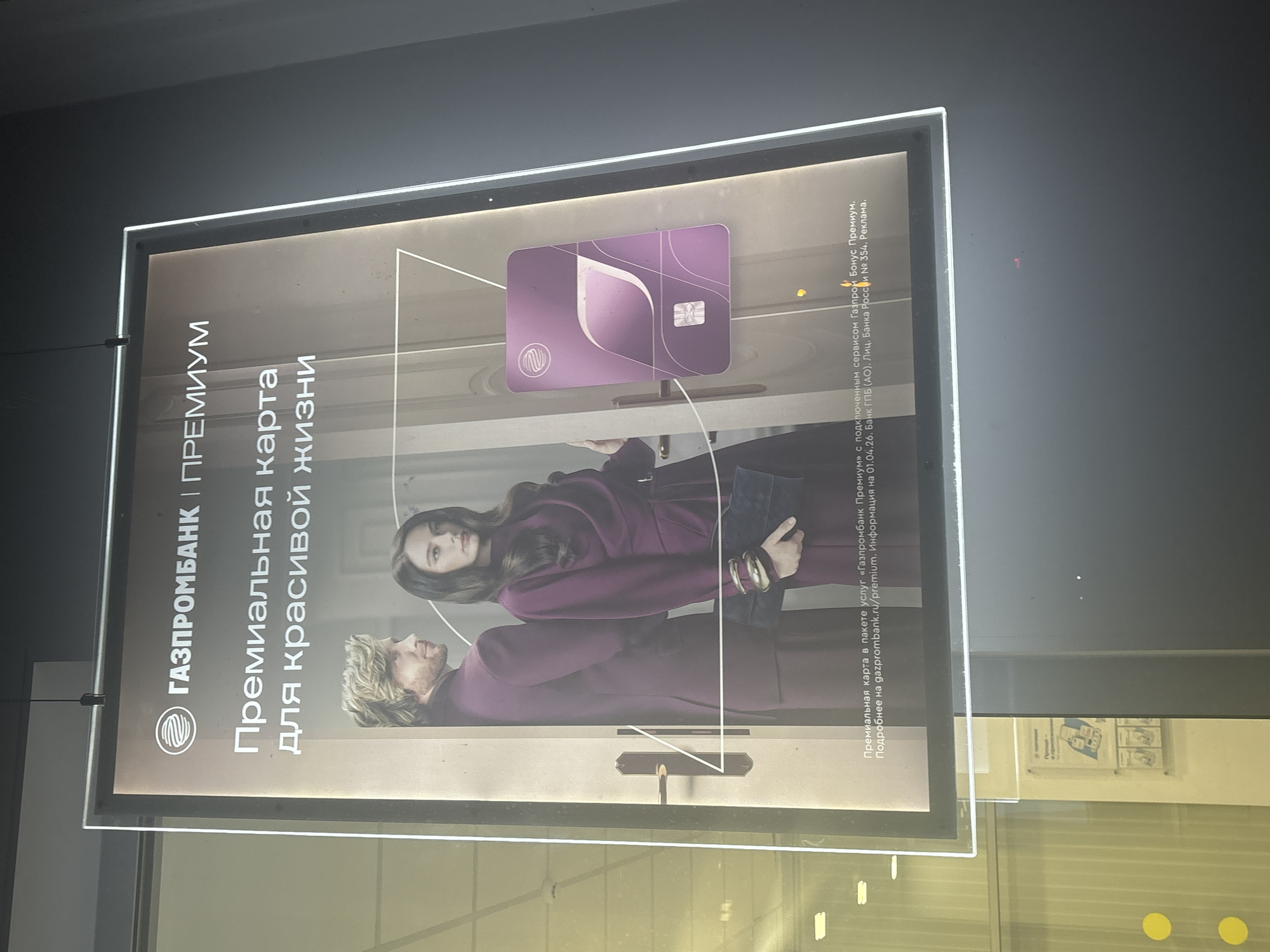 This is a horizontal, eye-level, close-up, slightly angled, outdoor photograph of an illuminated advertisement. The advertisement features a dark background and a central image of a man and a woman dressed in stylish, matching plum-colored attire, standing in what appears to be an entryway or doorway. They are both looking away from the camera. The man has blond, wavy hair, and the woman has dark, shoulder-length hair. To the right of the couple, a purple credit card with a stylized white logo is prominently displayed. Text in Russian is visible at the top of the advertisement, reading "ГАЗПРОМБАНК | ПРЕМИУМ," which translates to "Gazprombank | Premium." Below this, there is more Russian text advertising a premium card for a beautiful life. The bottom of the advertisement contains smaller text with details about the service, including a website address and information about the bank.
The advertisement is framed by a thin, white border and is suspended by thin black wires. The background outside the frame shows reflections of indoor lighting and a glass storefront, suggesting the advertisement is displayed in a public space, possibly a bank branch or a shopping mall, in Novosibirsk, Russia. The overall lighting is dim, with the advertisement being the brightest element. Yellow circular stickers are visible on the glass in the lower left portion of the image.