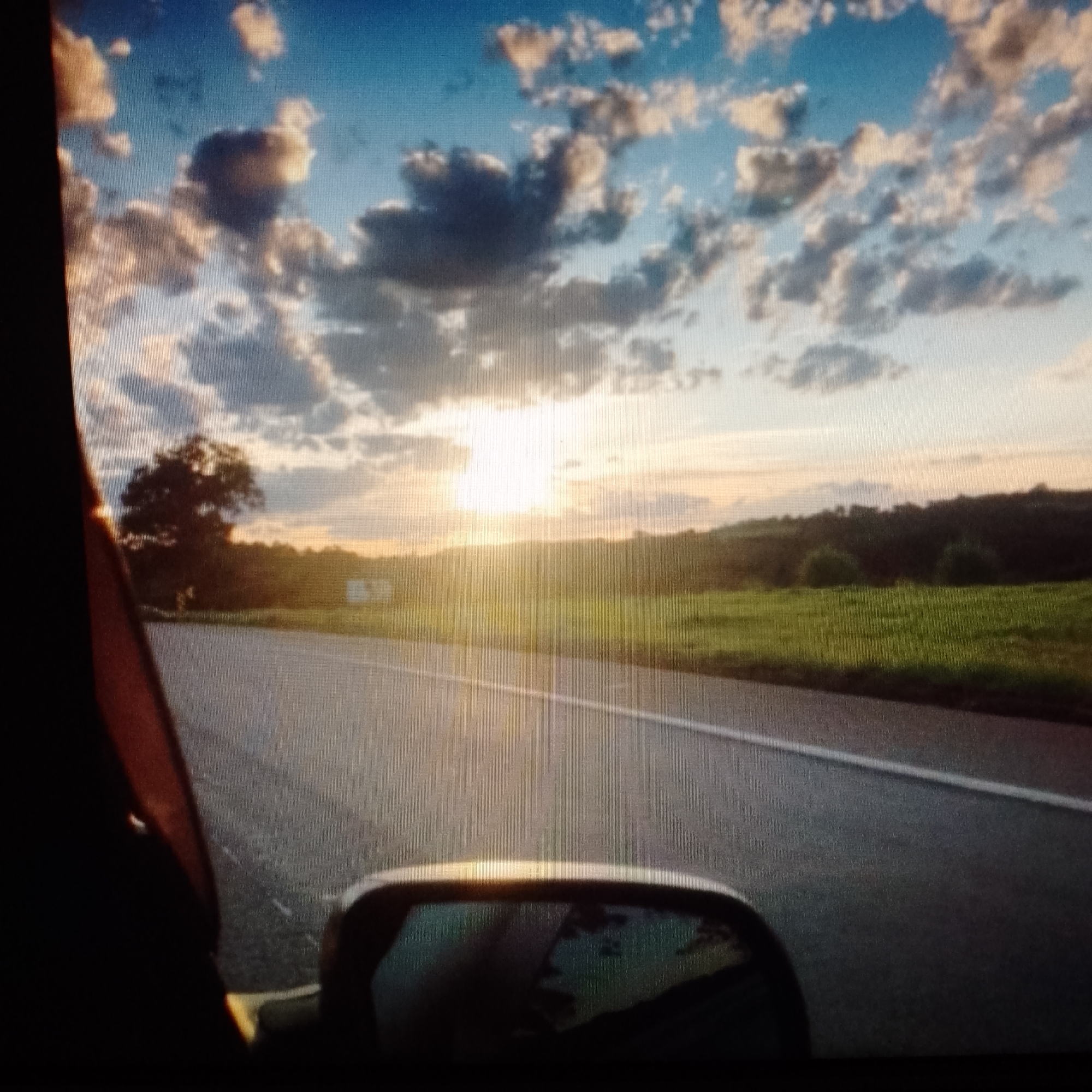 A view from inside a car, looking out a side mirror onto an asphalt road with a white line marking the lanes. Beyond the road, a grassy field extends to a treeline. The setting sun, partially obscured by clouds, casts a bright glow over the landscape. The location is rural, unidentified city or region.