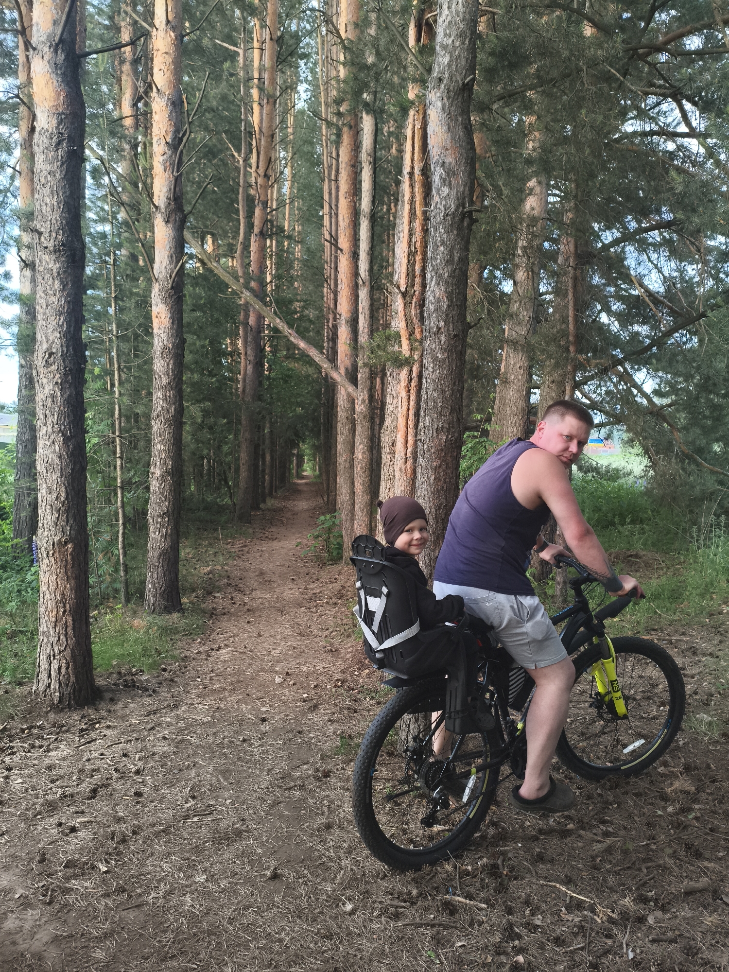 An adult male and a child are riding a bicycle on a dirt path in a pine forest. The man, wearing a dark blue tank top and grey shorts, is in the foreground, looking at the camera. He has tattoos on his arm. The child, wearing a brown hat and dark clothing, is seated in a black child seat on the back of the bicycle. The path winds through a dense stand of tall pine trees. The ground is covered with pine needles and cones. The lighting suggests it is daytime, possibly late afternoon. The scene appears to be in a natural, wooded area, likely a park or recreational trail. The location is identified as Русский Вожой, Russia, but this cannot be visually confirmed from the image itself.