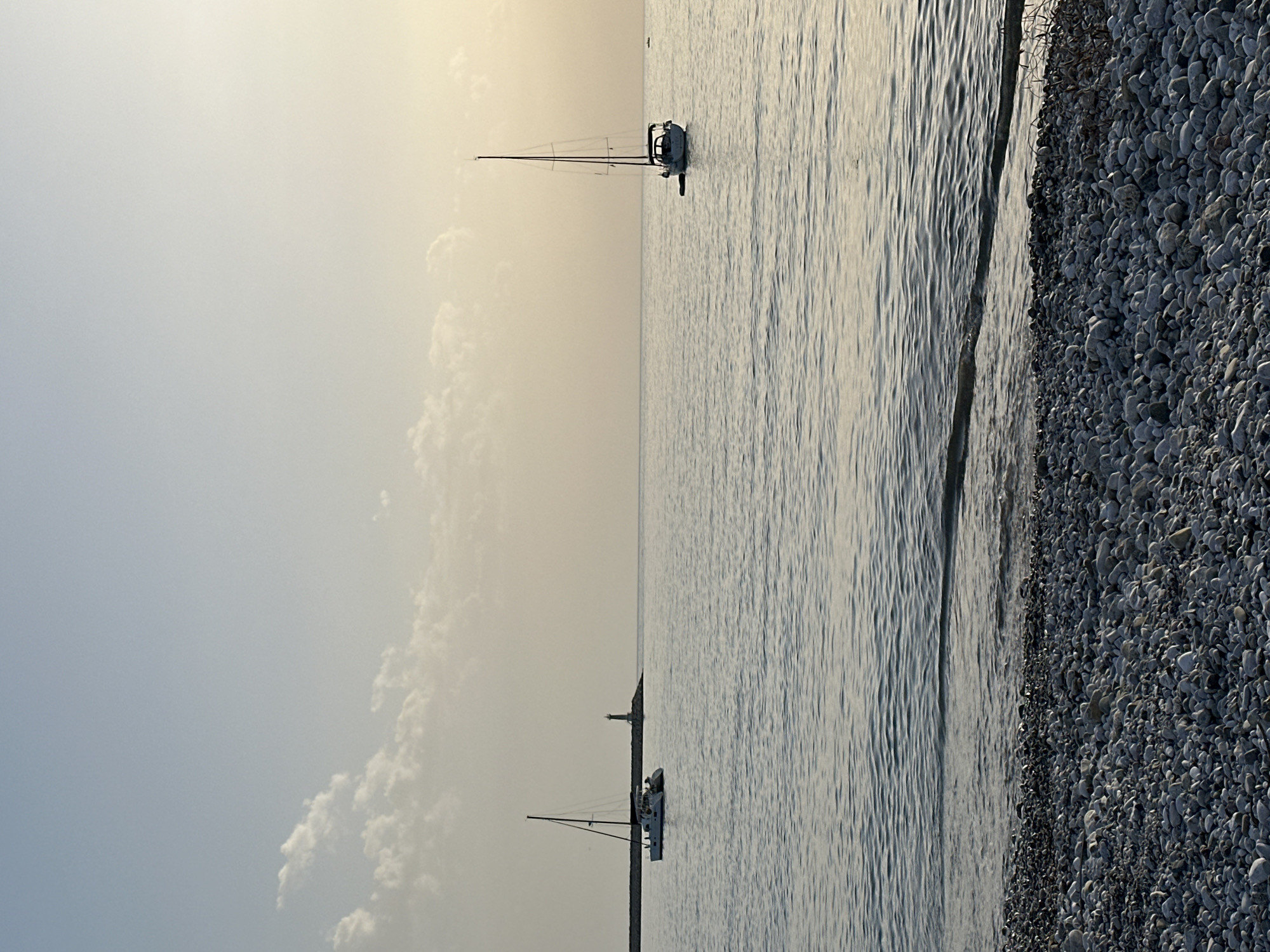 The image shows a seascape with a rocky beach on the right side. The water is calm and reflects the sky, which is hazy. Two sailboats are visible in the distance. The image appears to be taken at Šušanj, Montenegro due to its rocky beaches and calm waters. There are no people visible in the image.