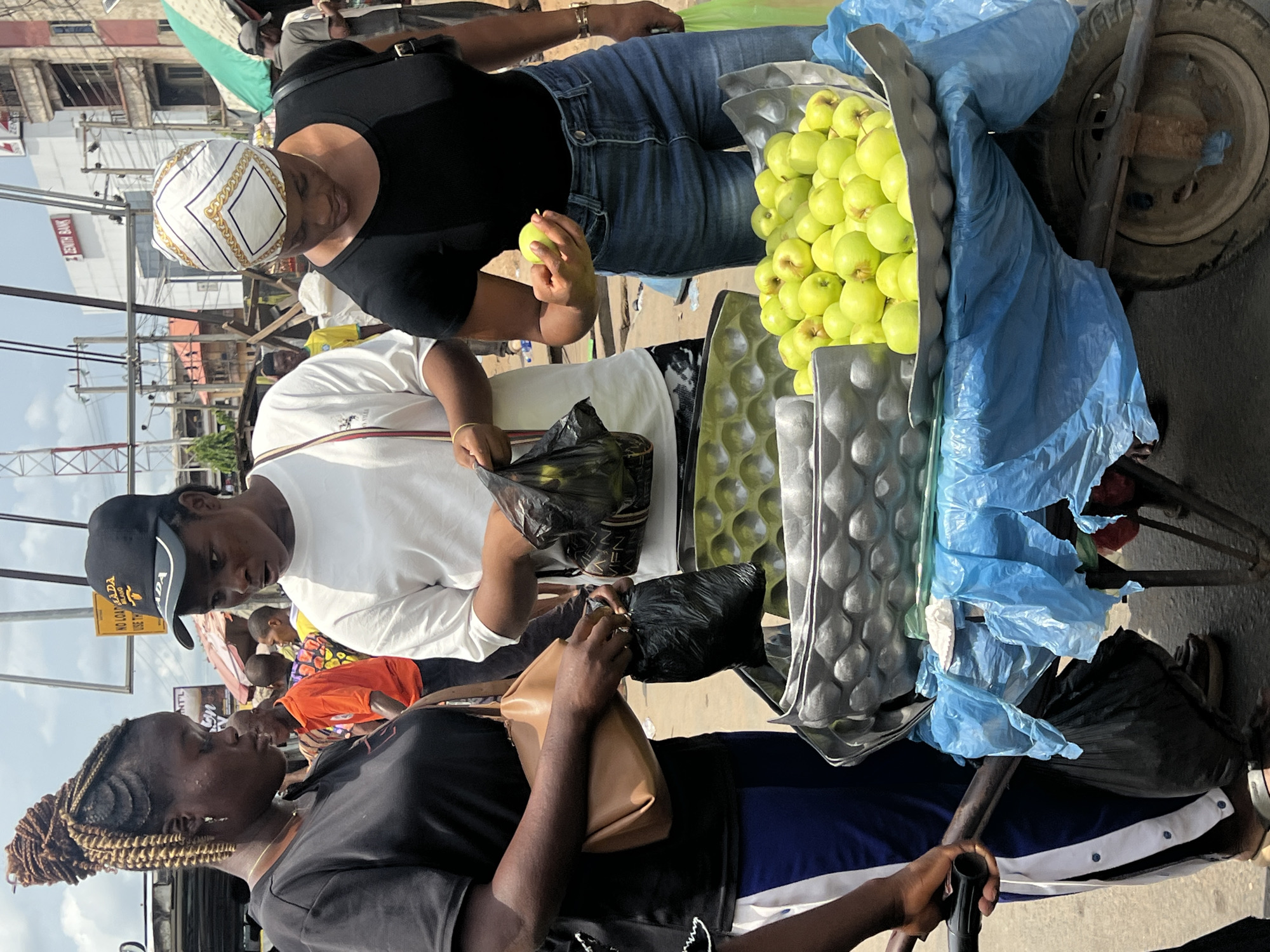 On a sunny day in what appears to be Onitsha, Nigeria, three women are interacting around a wheelbarrow laden with green apples. The wheelbarrow has two levels of apples separated by a gray packaging material and covered partially by a blue plastic sheet.
The woman closest to the viewer is wearing a black shirt and blue athletic pants with white stripes. She is holding a tan purse and the wheelbarrow handle. Another woman in a white t-shirt and a black cap is placing apples into a black plastic bag. She is looking at the woman in the black shirt.
The third woman, wearing a black shirt, blue jeans, and a white and gold headscarf, is holding an apple and looking at it. She is standing near the front of the wheelbarrow. The background includes a busy street scene with buildings and a Zenith Bank sign.