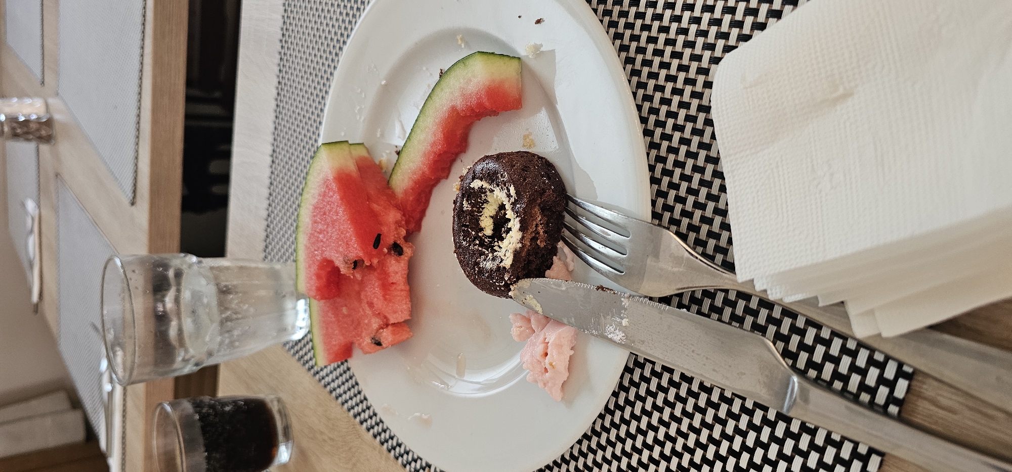 A close-up, slightly elevated shot captures a white plate on a black and white checkered placemat at a table, likely in a restaurant or hotel dining area in Supetar, Croatia. On the plate are remnants of a meal, including three slices of watermelon, one of which has bite marks, a small chocolate cake with a bite taken out of it, and a dollop of pink frosting or ice cream. A silver knife with a serrated edge rests to the left of the plate, and a silver fork is positioned to the right. A stack of white paper napkins is situated in the foreground, partially obscuring the lower portion of the silverware. In the background, two glasses are visible: one is a clear glass filled with ice and water, and the other, to the left, is a short, dark glass containing a dark beverage, possibly cola. The table surface is wooden, and part of a salt or pepper shaker is visible in the upper background. The lighting is natural and soft. There are no people in the frame.