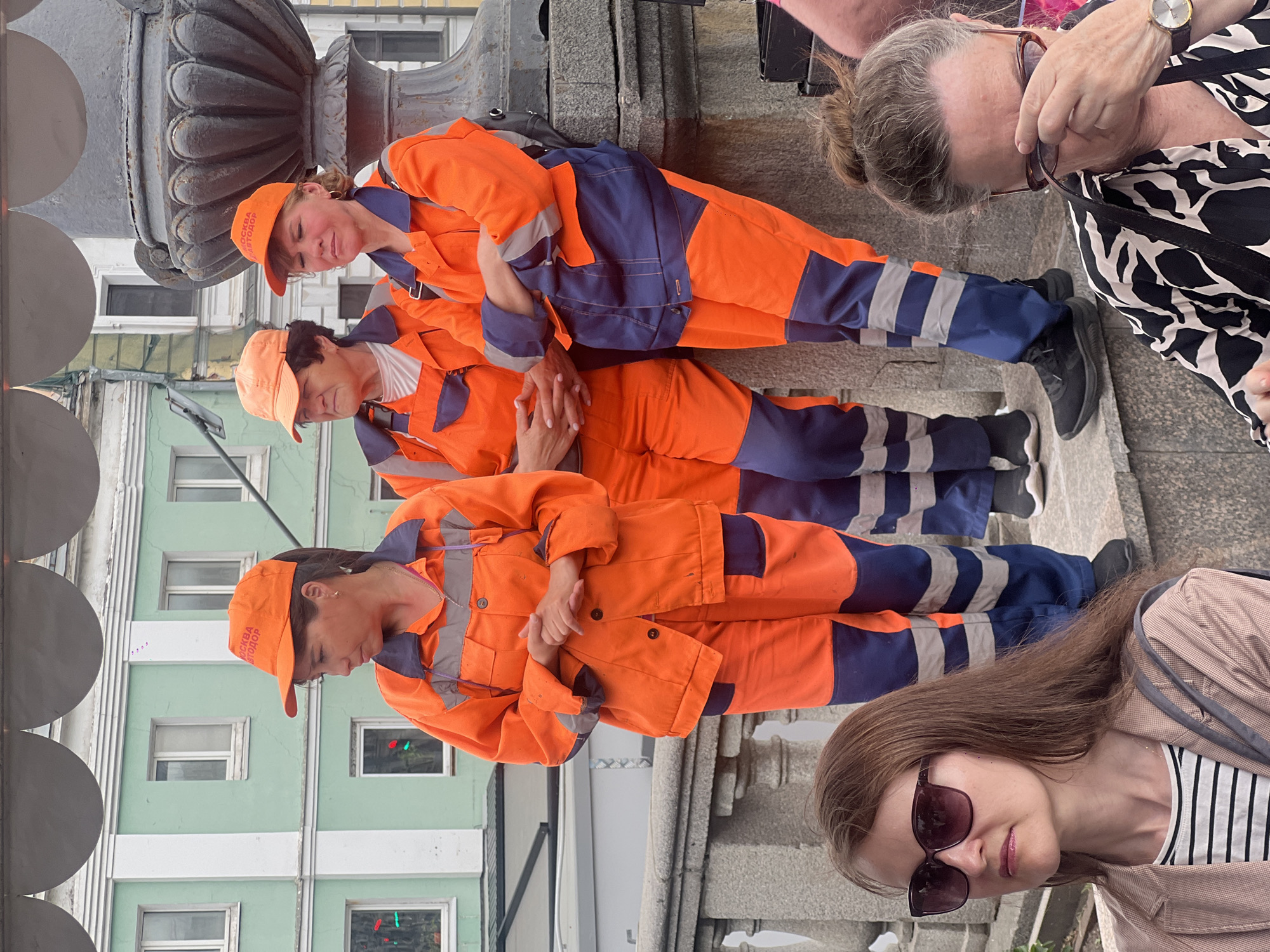 Three women in orange and blue work uniforms and orange caps are standing in a line. They are posed against a concrete structure with a decorative urn-like element behind them, and a building with windows is visible in the background. Two women are standing to the side, partially visible. The location is in the city of Unknown, Unknown.