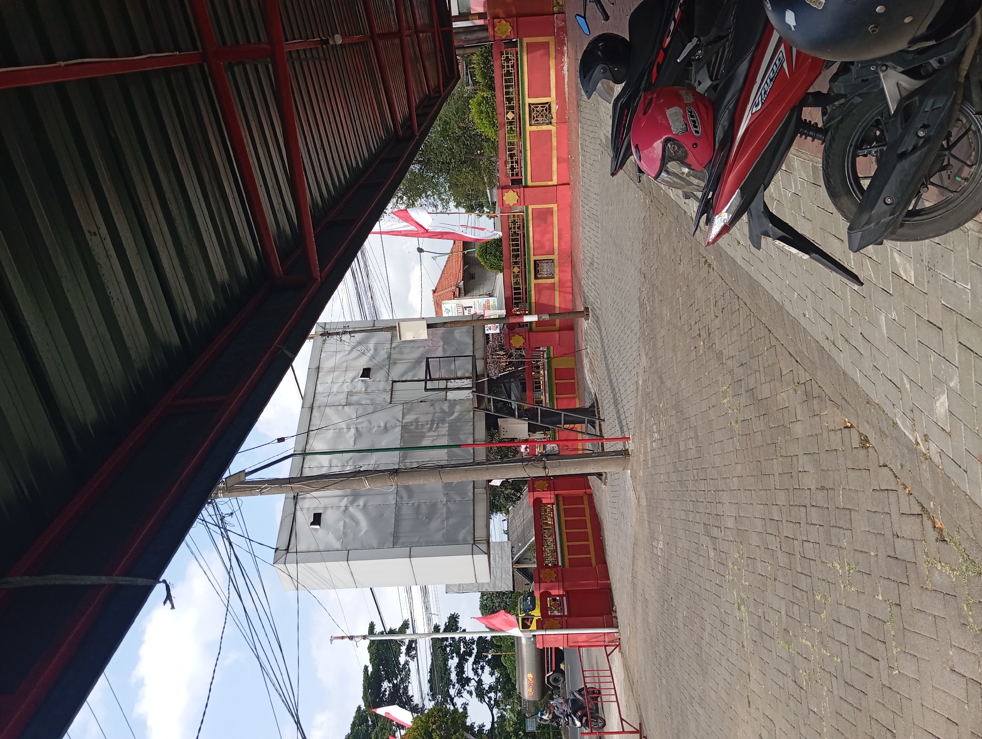 The image shows a street scene in Pandaan, Indonesia, likely near a commercial area or roadside establishment. In the foreground, two motorcycles are parked, one red and one black, with helmets placed on their seats. The ground is paved with interlocking gray bricks. A red and yellow patterned wall with decorative elements runs along the middle ground. Behind the wall, a large, modern, silver-colored structure with visible windows stands prominently. A metal staircase leads up to this structure. Indonesian flags are visible on flagpoles, one to the left of the silver structure and another on the right, near the wall. A yellow tanker truck is on the road to the left, alongside some trees and a building. Power lines crisscross the sky above. The scene appears to be outdoors under a clear sky with some clouds, and the area is covered by a corrugated metal roof supported by red beams, suggesting a sheltered waiting area or awning.