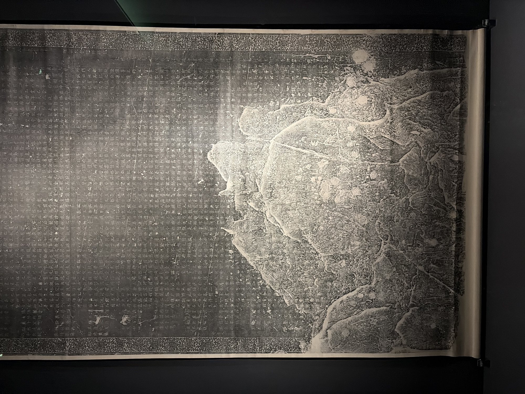 The image displays a large, dark calligraphy scroll with a textured, mountainous landscape depicted at the bottom. The scroll is unrolled and mounted on a dark background, suggesting it is an exhibit in a museum or gallery. The calligraphy is in Chinese characters and appears to be very old, with some characters faded and the paper showing signs of age. The mountainous imagery is rendered in a detailed, almost impressionistic style, with strong contrasts between light and shadow. The overall impression is one of historical and artistic significance.

This artifact is likely displayed at the National Museum of Korea, located in Yongsan-gu, Seoul, South Korea. The museum houses a vast collection of Korean art and history, including ancient artifacts and traditional calligraphy.