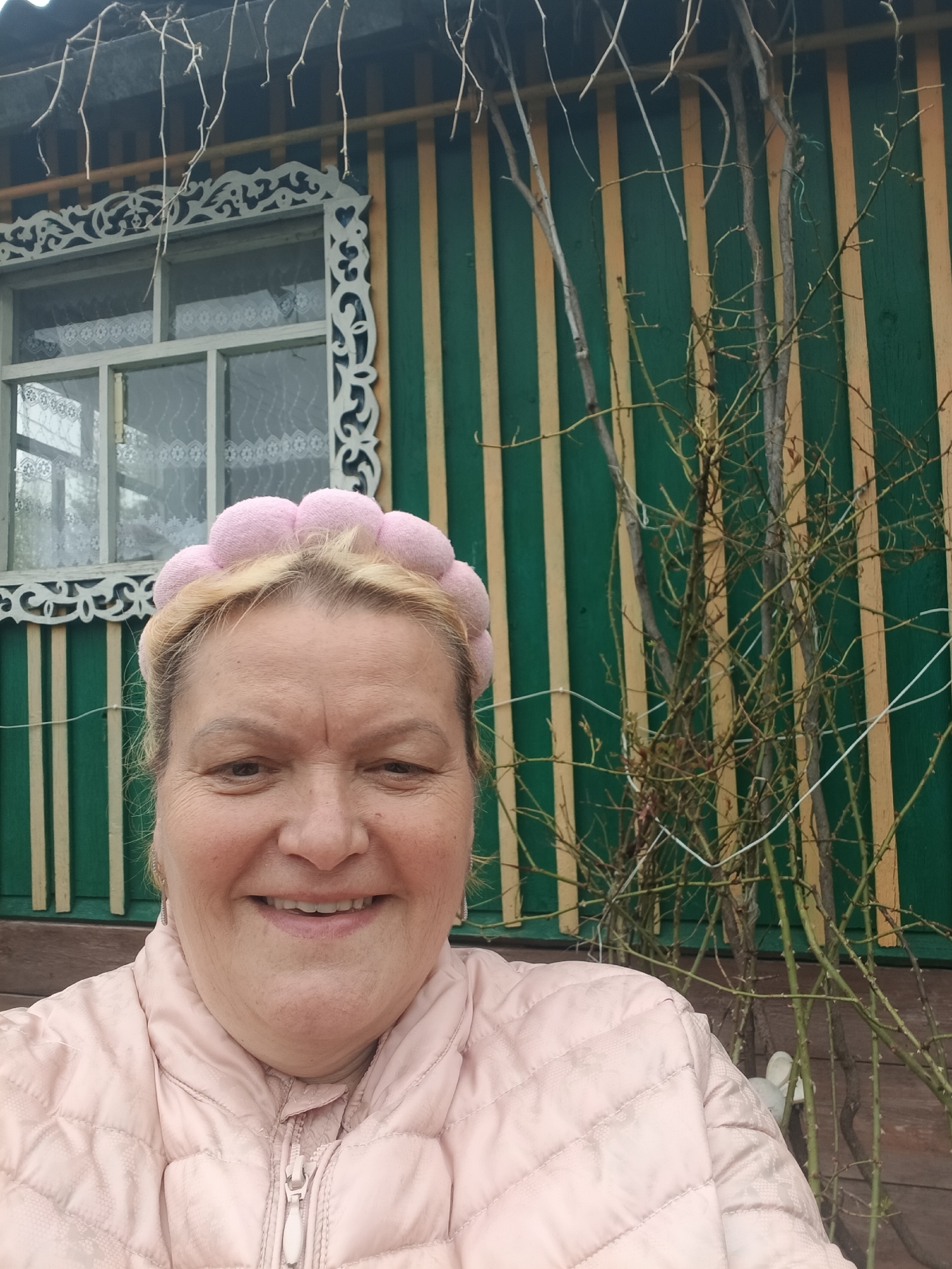 The image depicts a smiling, mature woman wearing a light pink quilted jacket and a pink headband. She appears to be posing for a selfie. Behind her, a building with green and yellow vertical paneling is visible. There is a window with decorative white trim to the left of the woman. In front of the building and to the right of the woman are bare branches, possibly from a rose bush, partially obscuring the wall. The location is Красный Маяк, Russia.