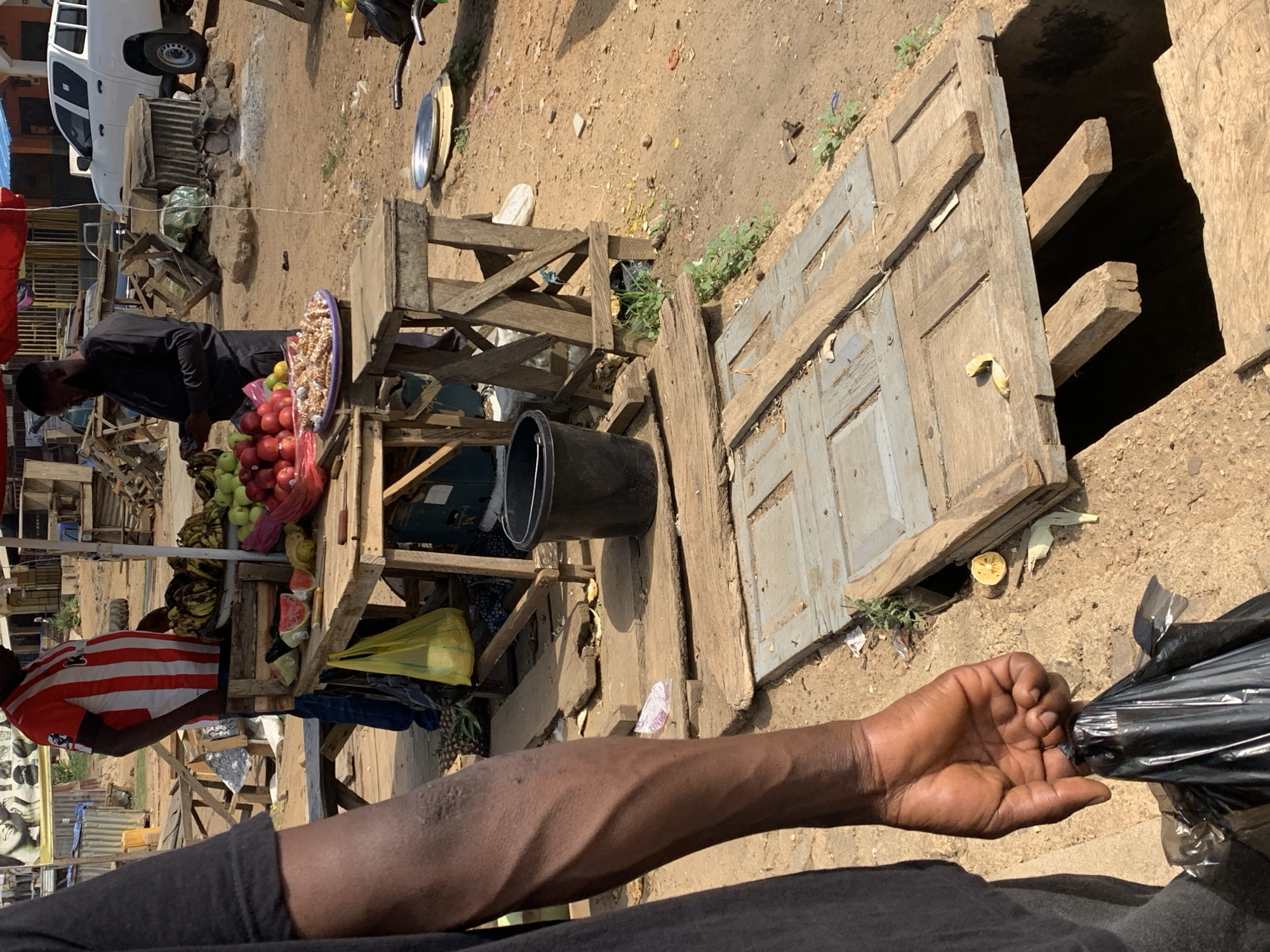 The image depicts a marketplace in Rafin Par, Nigeria. A vendor in a striped shirt stands behind a wooden fruit stand displaying apples, bananas, and watermelon. Another man in dark clothing browses the fruit. Beside the stand, there is a makeshift wooden door leading to a hole in the ground. A person is holding a black plastic bag, obscuring the view of the scene. The ground is dry and dusty. In the background, a white truck is parked near the market stalls.