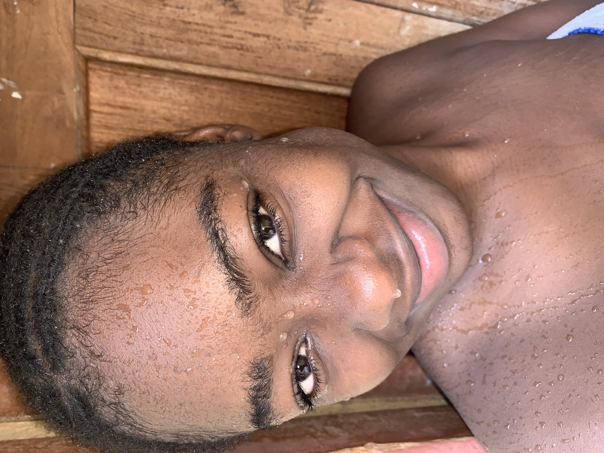 A close-up, tilted image shows a Black child with short, curly black hair and wet skin. Water droplets are visible on their face and upper body. The child is smiling slightly. In the background are wooden planks. There are no indications of a specific location in Rafin Par, Nigeria.