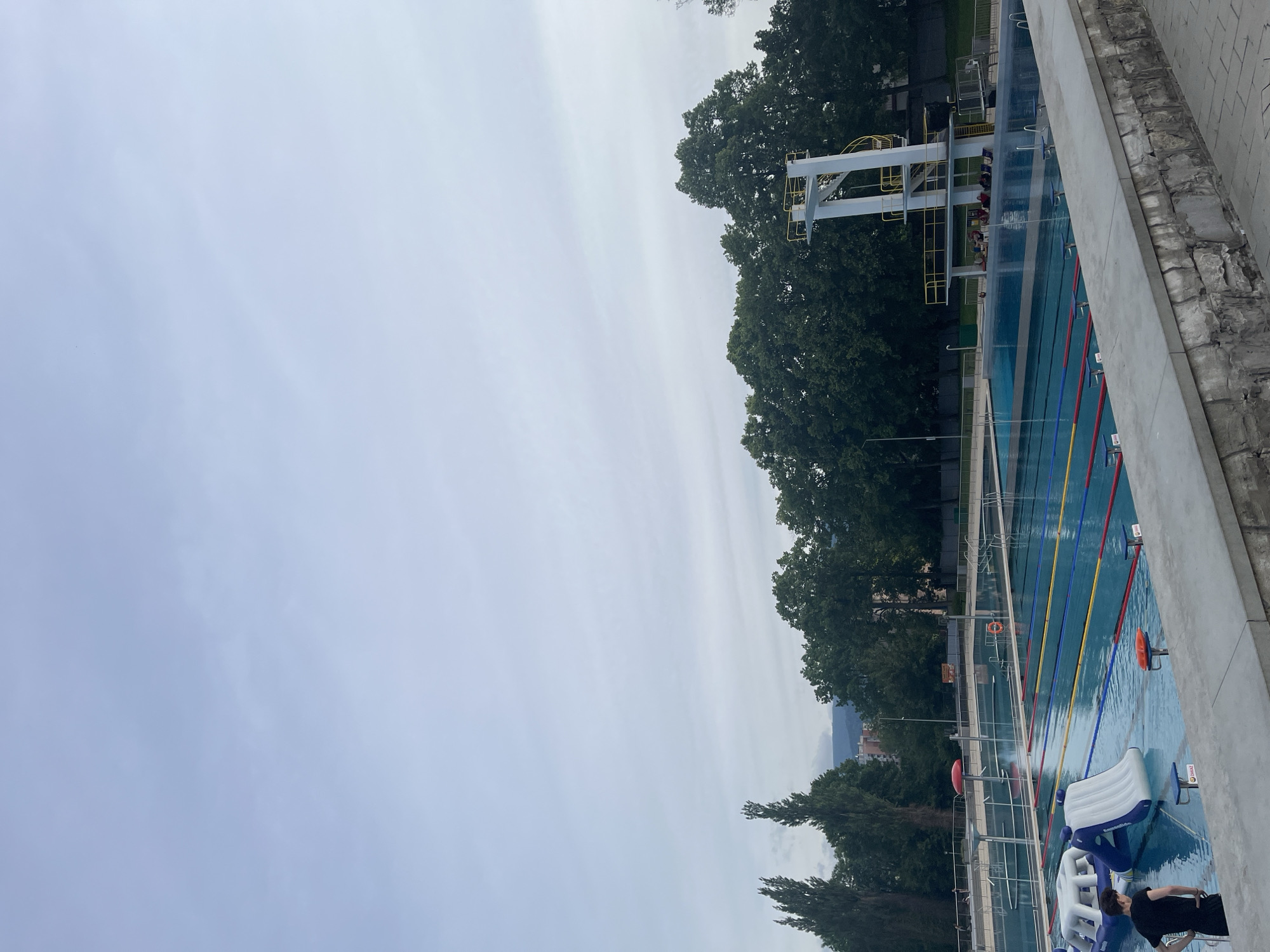 The image captures an outdoor swimming pool complex under an overcast sky. The main subject is a large, rectangular swimming pool with several lanes clearly marked by colorful ropes in red, blue, and yellow. Blue-topped starting blocks are visible along the edge of the pool.
On the far side of the pool, a tall, multi-level diving platform with yellow railings stands against a backdrop of dense, dark green trees. In the distant background, subtle outlines of hills or mountains and possibly buildings are visible.
Within the pool's shallower section, there are several white and blue inflatable water play structures, one of which is clearly branded "Wibit". A person, dressed in a dark shirt and shorts, is partially visible in the bottom right corner, standing next to one of these inflatables. Other indistinct figures can be seen near the diving platform. Orange and red life preservers are also present along the poolside.
The immediate foreground on the right consists of a textured concrete wall. The sky is uniformly grey and overcast, suggesting a cloudy day without direct sunlight. Visible text includes "ZAAKZ" on some starting blocks/lane markers and "Wibit" on an inflatable structure. The scene suggests a public or recreational swimming facility in active use or prepared for activity.