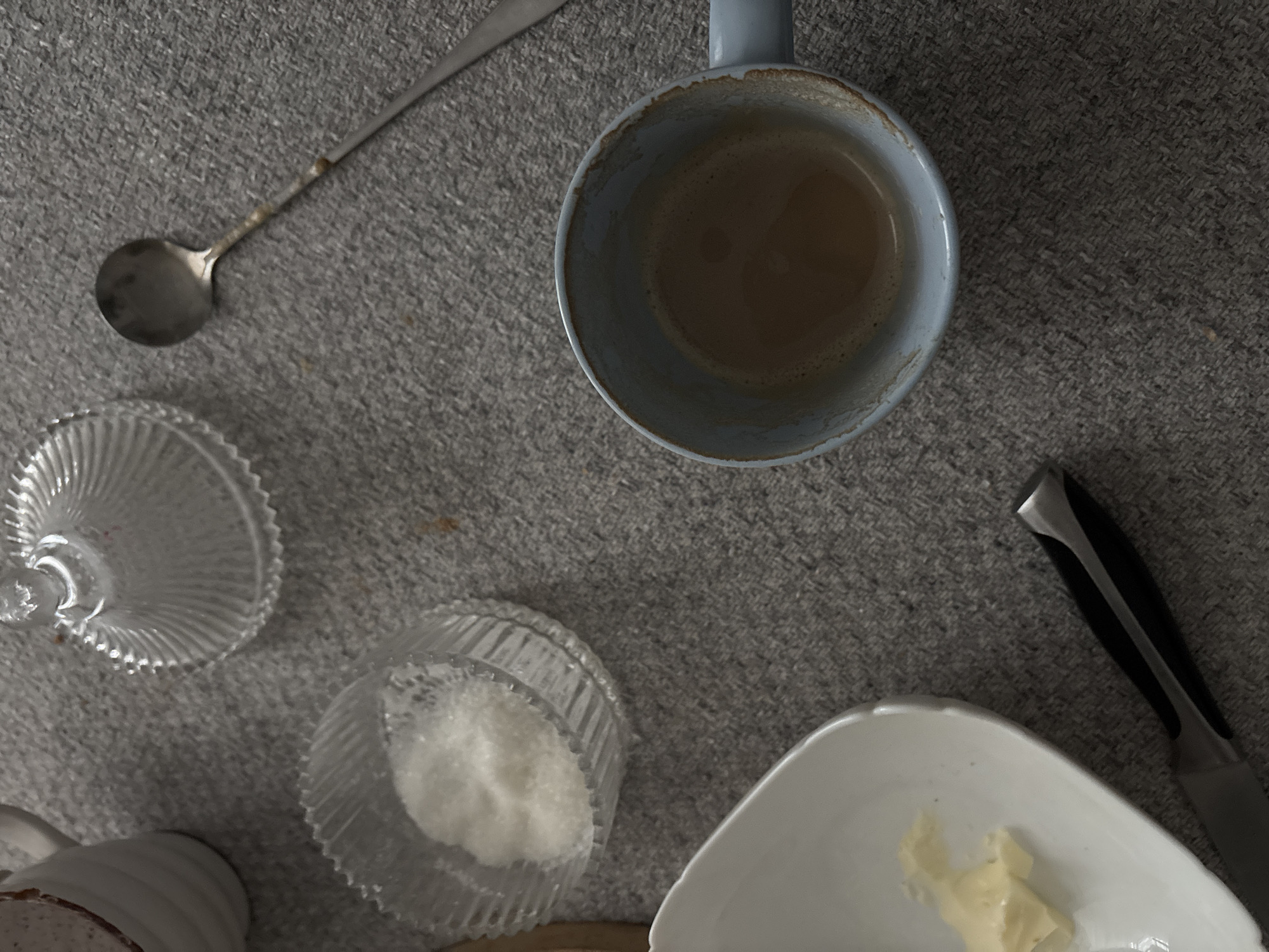 The image depicts a flat lay arrangement of objects on a textured gray surface. A light blue mug containing coffee sits near the center, with a tarnished silver spoon to its upper left. A fluted glass sugar bowl with a lid sits to the left, near a small glass bowl containing white sugar. A white, irregularly shaped dish with butter is in the lower right corner, adjacent to a knife with a black handle. No people or indications of a specific location, such as Zhlobin, Belarus, are apparent.