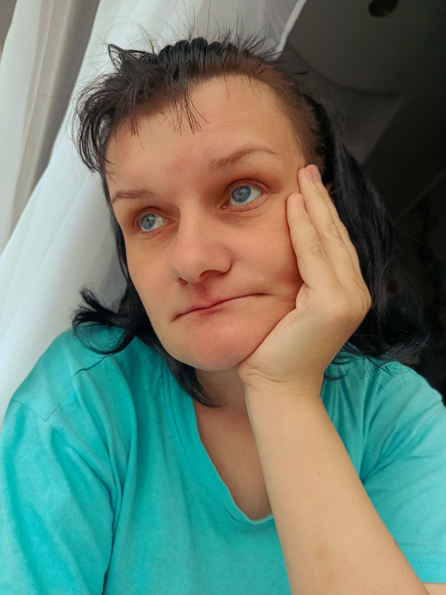 A woman with blue eyes and dark hair is shown in a close-up shot, her right hand supporting her chin. She is wearing a teal t-shirt. A white curtain is visible to her left. The scene appears to be indoors. There is no information available to determine the location as Фоки, Russia.