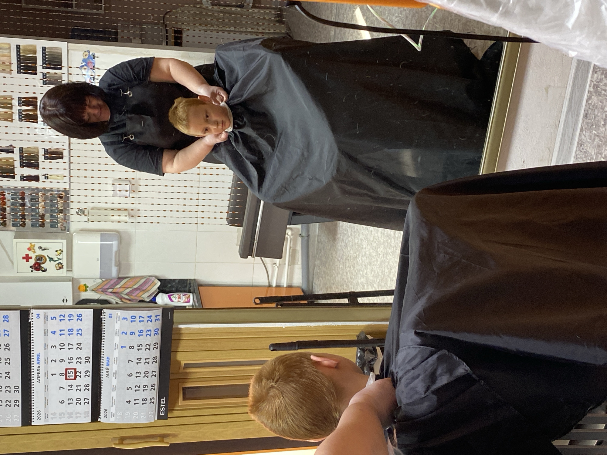 The image shows a woman with dark hair cutting a child's hair in what appears to be a hair salon. The woman is wearing a black smock and is holding the child, who is draped in a black cape. The child has blonde hair. The reflection in a mirror shows another child also getting their hair cut. A calendar on the wall shows the months of April and May 2026. Shelves with hair color samples are visible in the background. It is not possible to determine the exact location in Noviy, Russia from the image alone.