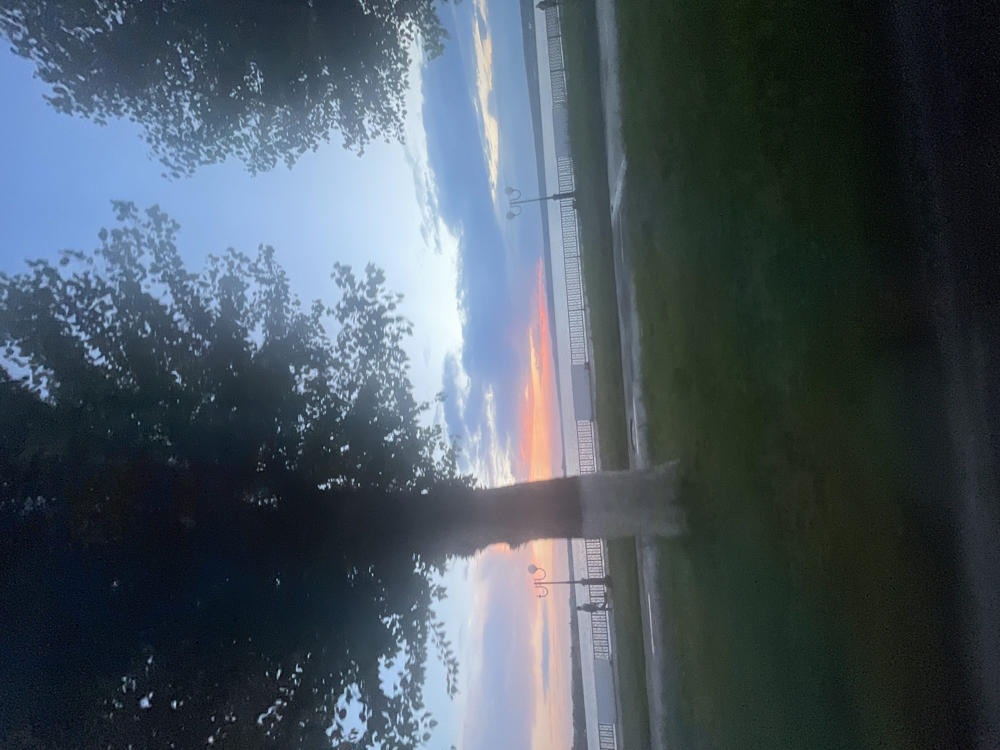 This image depicts a scenic view of the Votkinsk reservoir in Votkinsk, Russia, during sunset. The sky is painted with hues of blue, pink, and orange, transitioning into a clear blue overhead. Silhouetted trees frame the scene, their branches casting shadows on the foreground. A paved walkway runs along the reservoir, bordered by a decorative metal fence. A lamppost stands on the walkway, illuminated by the fading light. A lone individual is visible walking on the pathway. The foreground features a grassy area.